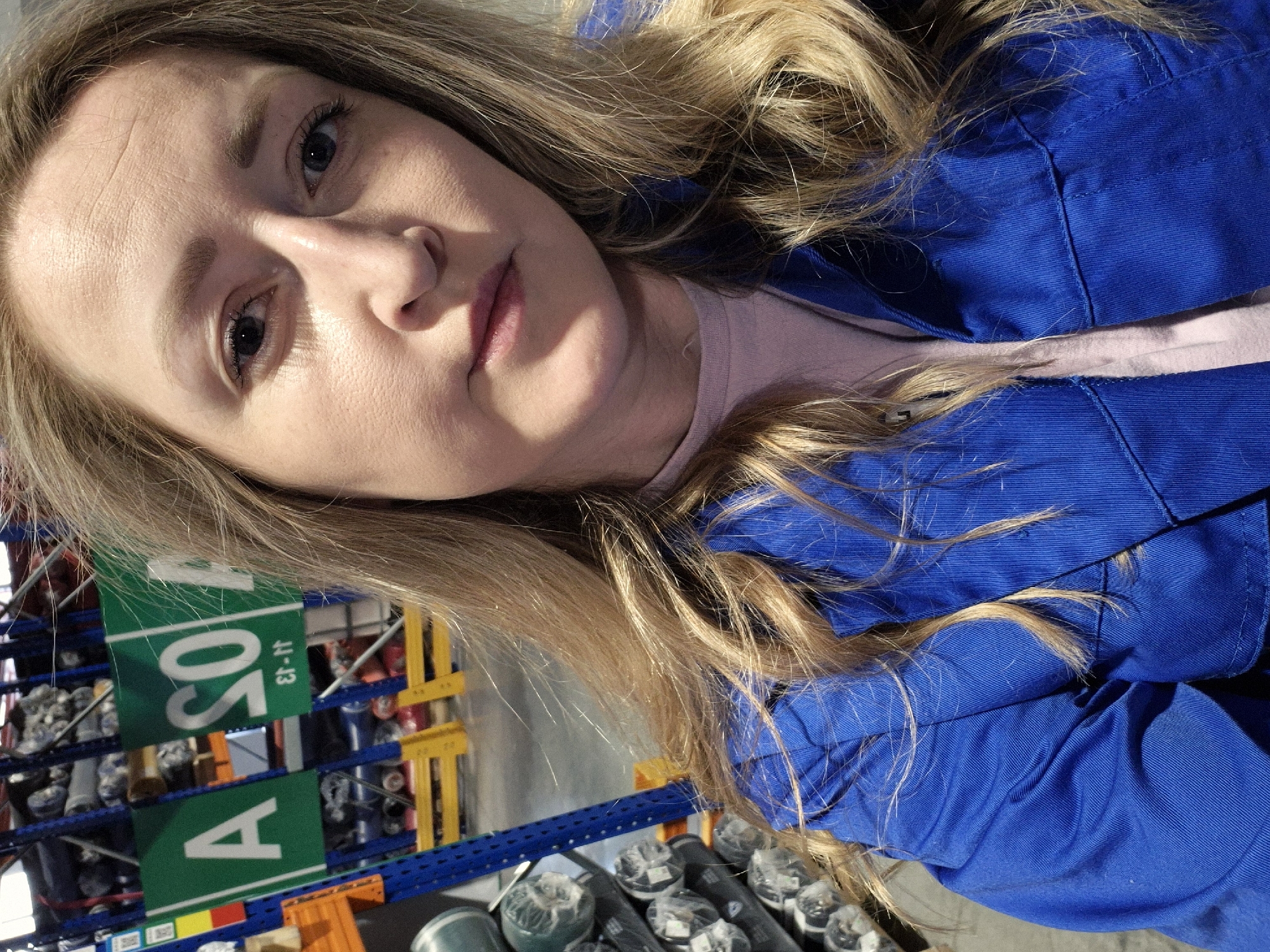 A selfie shows a fair-skinned woman with light blonde hair, wearing a light pink shirt under a bright blue collared jacket or uniform, in what appears to be a warehouse or storage facility. Shelving units with various rolled items are visible in the background. A sign on the shelf reads "A 501 11-13".
The location cannot be specifically identified as Chaikovsky, Russia from the visible content of the image.