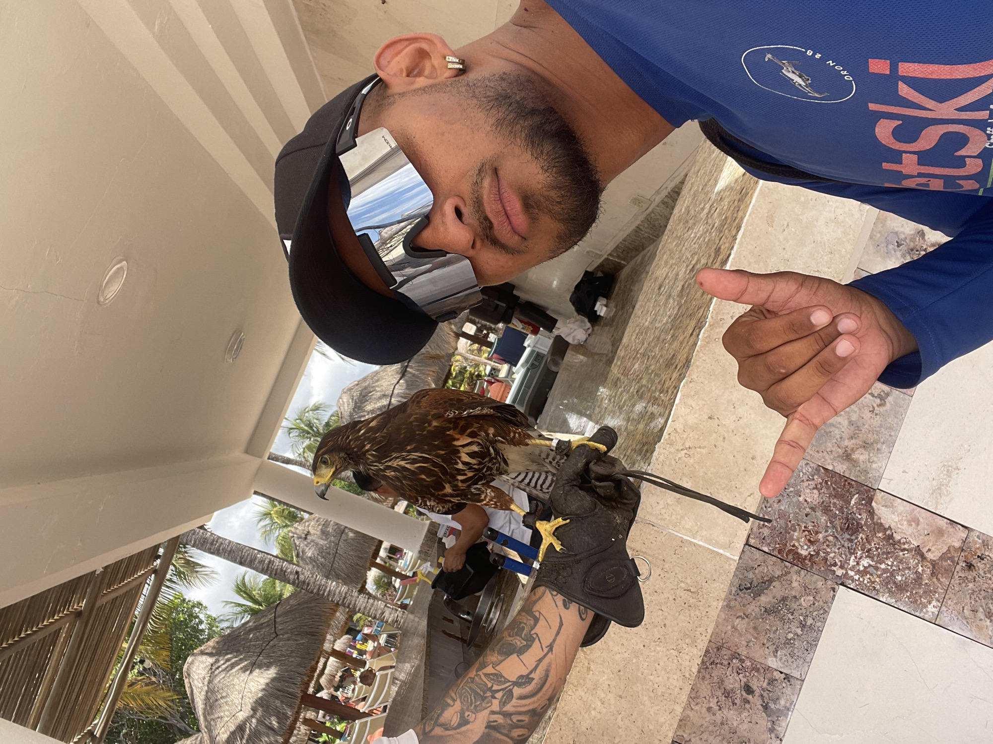A man is holding a Harris's hawk in Playa del Carmen, Mexico. The man is wearing a blue shirt, a black baseball cap, and mirrored sunglasses. He has a gold earring and a beard. His arm has tattoos. The Harris's hawk is perched on a leather glove. The background shows thatched-roof palapas and beach chairs, indicating a resort or tourist area. The floor is a marble pattern.