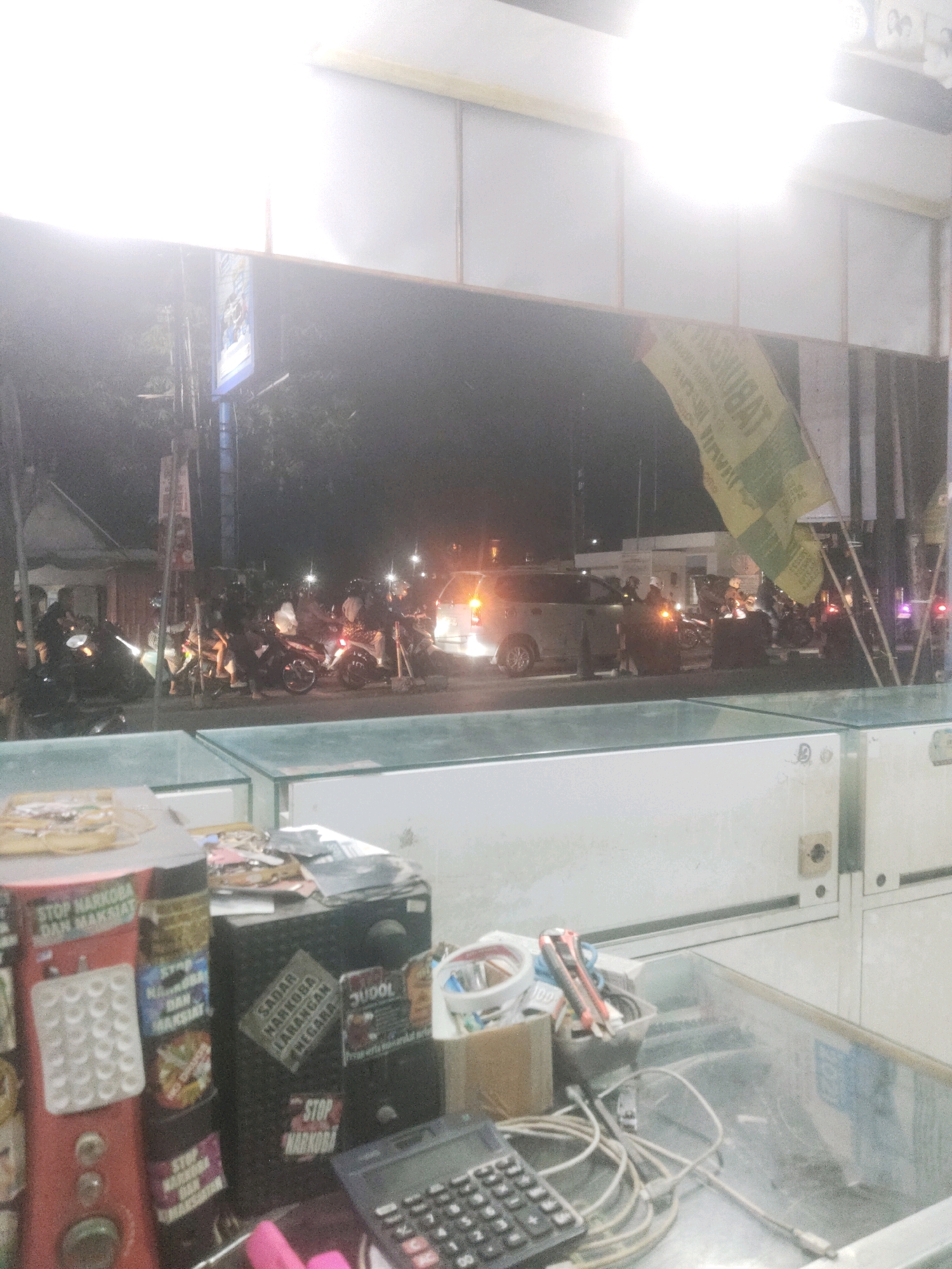In Sragen, Indonesia, at night, a street scene unfolds. A variety of motorcycles are parked on the roadside, some with riders visible. A silver SUV is also present, its taillights illuminated. In the foreground, a glass display case reveals various items, including a calculator, spools of wire, and what appears to be a small box of supplies. Several signs with anti-drug messages are also visible. One sign prominently reads "STOP NARKOBA DAN MAKSIMAT". Another, on a black speaker, says "SADAR NARKOBA LARANG NEGARA". A yellow and green flag with text is draped over a pole, partially obscuring the background. The overall lighting suggests a dimly lit environment, typical of an evening street in a city.