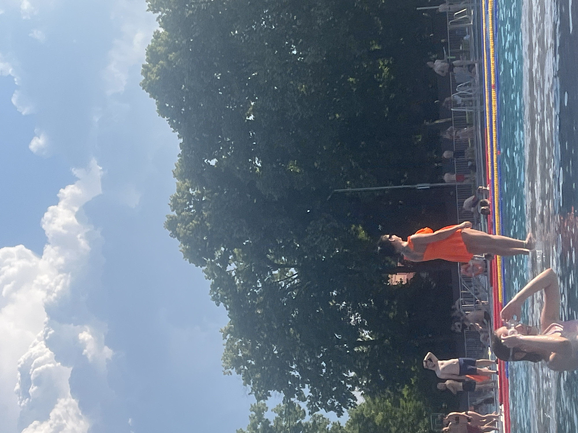 A sunny day at an outdoor swimming pool features several people engaged in various activities. In the foreground, a young woman wearing a pink swimsuit and swimming goggles is in the water, adjusting her goggles. In the middle ground, a woman in a bright orange one-piece swimsuit stands at the edge of the pool, facing away from the camera. Behind her, several men are visible. One man in blue shorts stands with his hand on his forehead as if shielding his eyes from the sun. Other people are scattered around the pool area, some sitting, some standing. The pool itself has distinct lanes marked by buoys in red, yellow, and blue. Beyond the pool, lush green trees form a dense backdrop, with a clear blue sky and fluffy white clouds above. The overall scene suggests a recreational setting on a warm day.