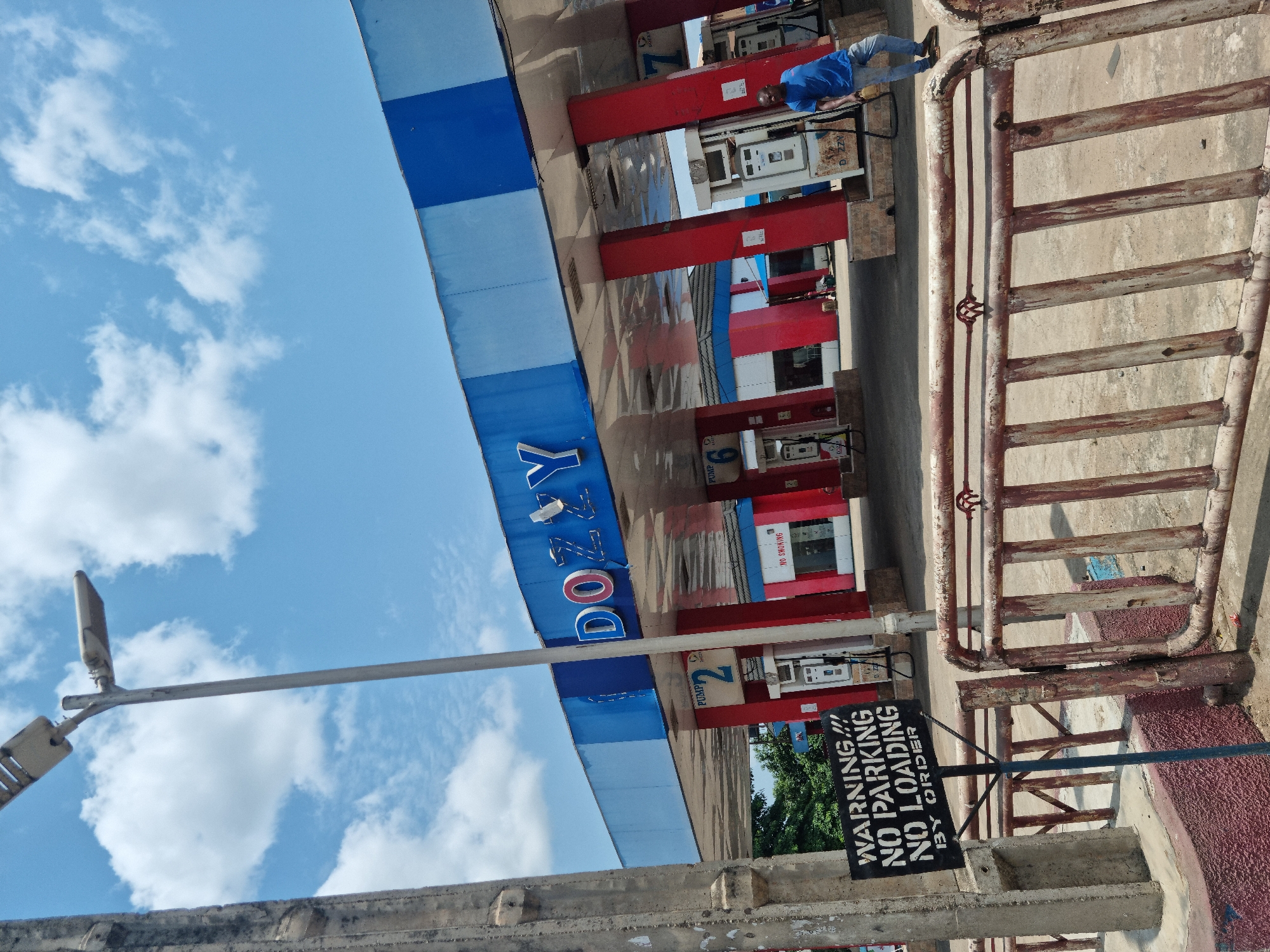 This daytime image, captured in Onitsha, Nigeria, depicts an active Dozzy gas station under a bright blue sky with scattered white clouds. The central feature is the station's canopy, prominently displaying the brand name "DOZZY" in white and red letters against a blue and white striped background.
Beneath the canopy, several fuel pumps are visible, each featuring red support structures and white dispensing units. Labels such as "PUMP 1," "PUMP 2," and "PUMP 6" are discernible on the pumps. A smaller sign on one pump explicitly states "NO SMOKING." A male attendant, dressed in a blue shirt and light-colored trousers, stands near the pumps, indicating ongoing service at the station.
The ground area around the pumps appears to be concrete, with some reddish-brown sections. To the right of the fuel pumps, a weathered, rusty metal barrier composed of horizontal bars separates the station area. In front of this barrier, a black sign with white text clearly reads: "WARNING!!! NO PARKING NO LOADING BY ORDER."
On the left side of the frame, a tall grey pole supports a street light fixture, extending upwards. A low concrete barrier is visible in the immediate foreground, partially obscuring the base of the pole. The polished surfaces of the pumps and the canopy's underside reflect elements of the sky and surrounding structures, adding depth to the scene of this functional gas station.