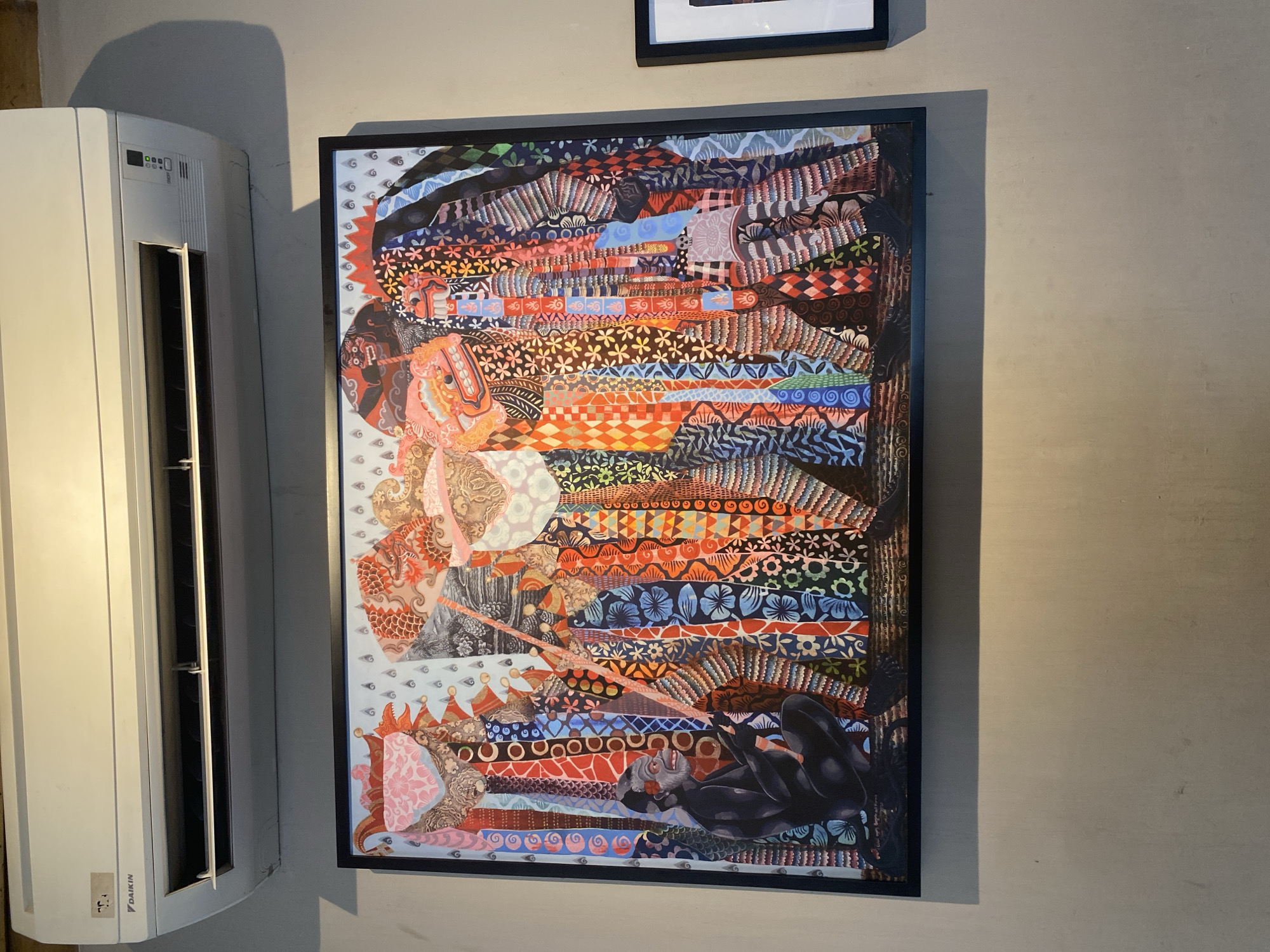 A photograph of an abstract painting with black frame hanging on a light grey wall next to a white air conditioner, taken in Mengwi, Indonesia. The painting appears to be a mixed media artwork with motifs and patterns resembling textiles, with figures or objects placed within the layers of pattern. The dominant colors are orange, red, blue, and black. At the bottom right, a black figure, perhaps a reclining person or animal, is visible. The wall has a neutral tone, and the air conditioner is a modern, wall-mounted unit. Another framed picture, only partially visible, hangs above the painting.
