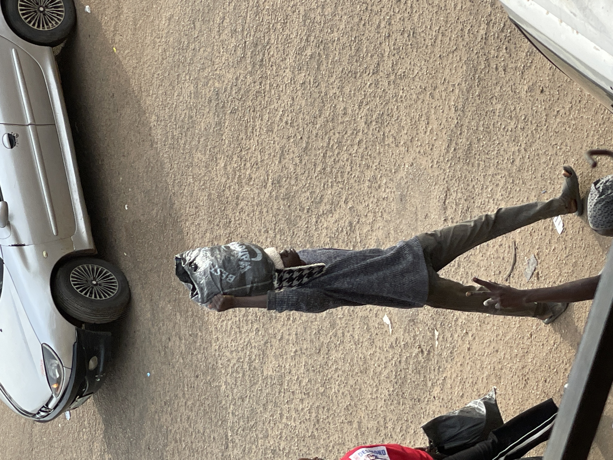 A medium shot shows a person from the side, walking on a dusty ground, carrying a large black plastic bag on their head. The person is wearing a dark grey sweater, dark grey pants, and sandals. A checkered scarf is visible around their neck. To the left of the person, another person in a red shirt and black pants with white stripes is walking away from the camera. In the background, a silver car is parked. The ground is covered in gravel and dirt, with scattered debris. The lighting suggests it is daytime, with shadows cast on the ground. The scene appears to be in an outdoor, urban environment.