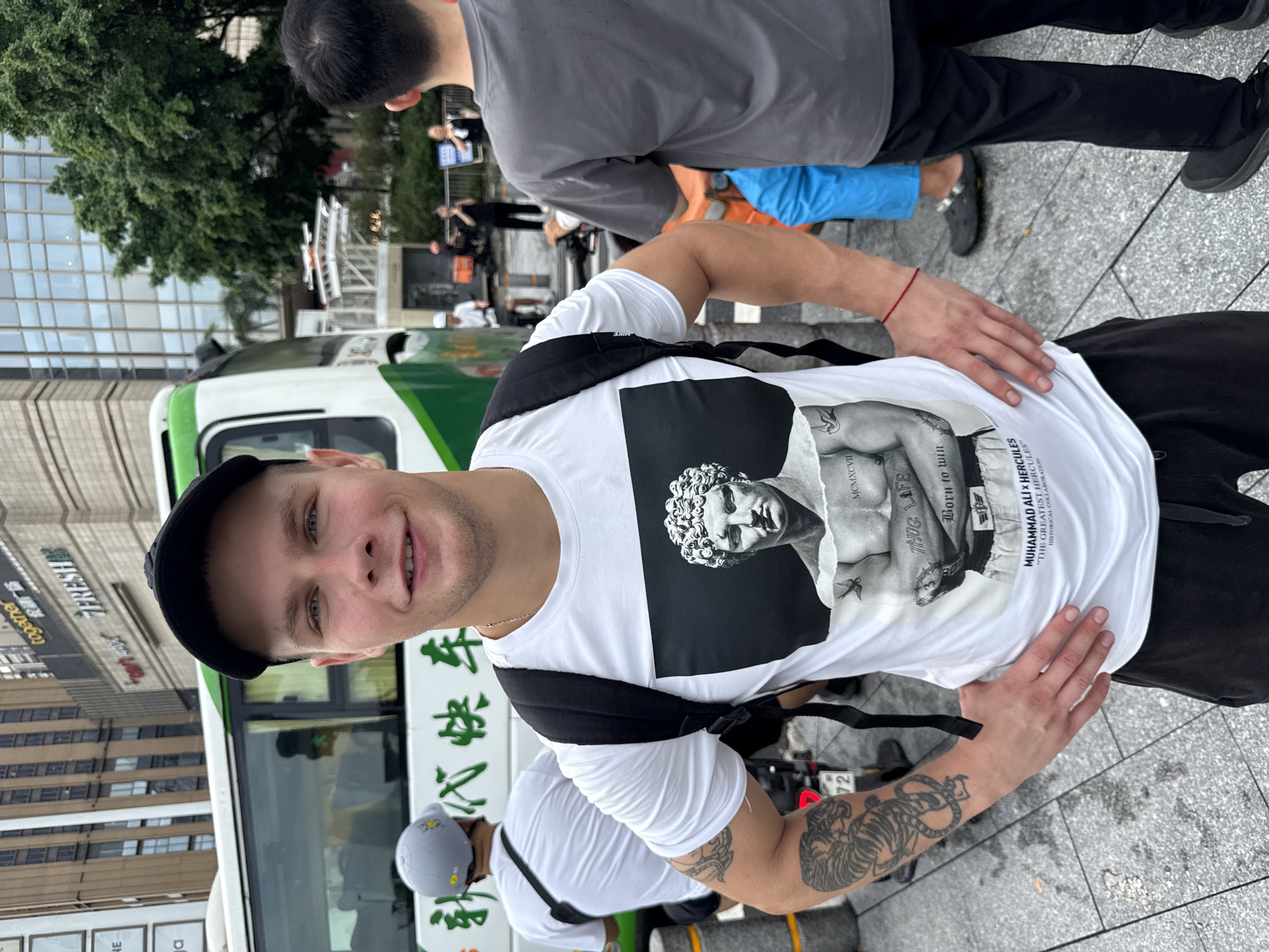 A young adult male stands in the foreground, facing the camera. He has short blonde hair and is wearing a black baseball cap. His eyes are light blue, and he has a slight smile. He is dressed in a white t-shirt with a graphic print of a statue of Hercules on it. The text on the shirt reads "MUHAMMAD ALI X HERCULES THE GREATEST HEROES" and "BORN TO WIN". He has a black backpack on. His left arm is visible and has a tattoo of a tiger and a snake on it. He is wearing black shorts. He is standing on a grey paved surface, possibly a sidewalk. In the background, a white and green bus is visible with Chinese characters on its side. Further back, there are buildings and trees. Another person is visible in the background, wearing a grey t-shirt and black pants. The scene appears to be in an urban environment, potentially in Haizhu District, China, given the presence of Chinese text on the bus.