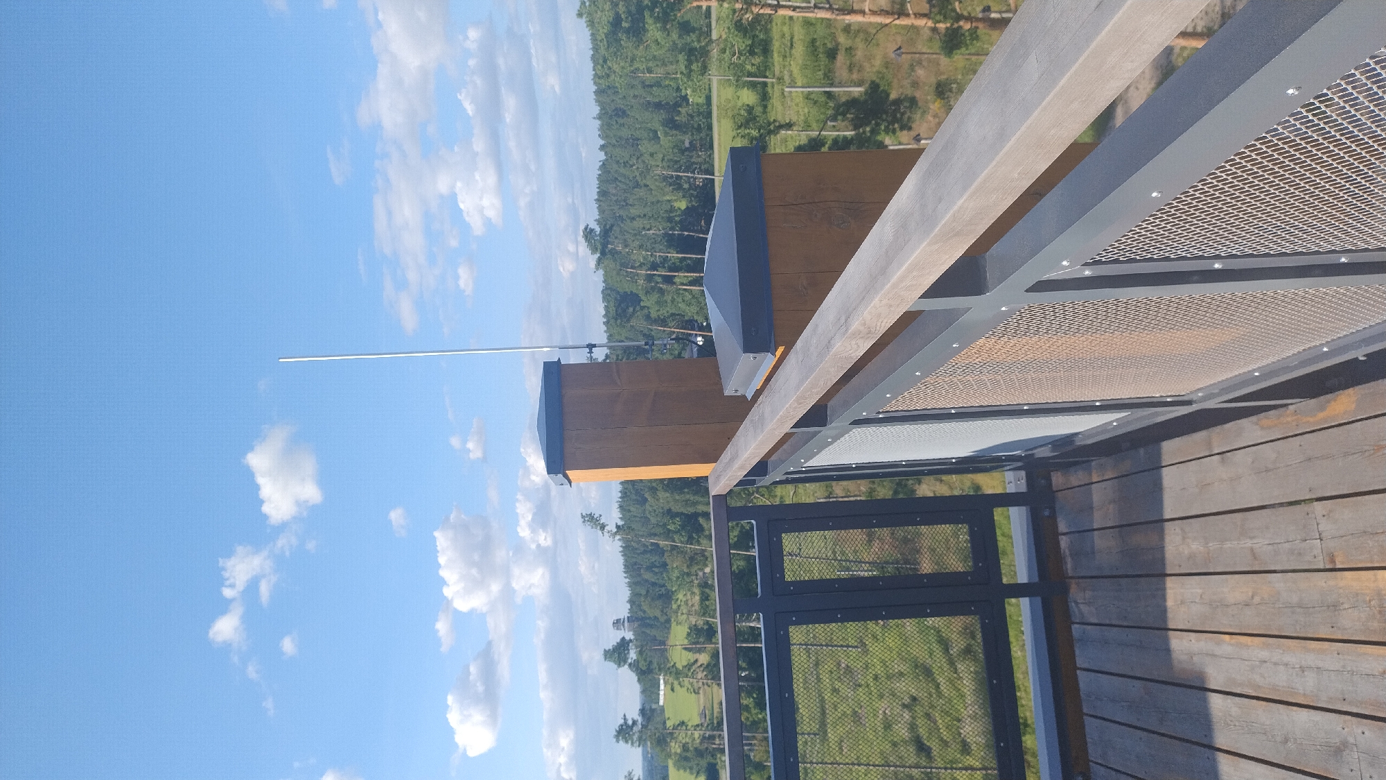 The image shows a view from a wooden observation deck with metal railings. The deck is made of weathered wooden planks and has dark grey metal railings with mesh infills. Two wooden support posts with dark grey caps are visible on the deck, one of which has a tall, thin antenna attached. In the background, there is a scenic landscape of green fields, trees, and a forest under a bright blue sky with scattered white clouds. In the distance, a structure resembling a tower is visible. This scene is located at the Tērvete Nature Park in Tērvete, Latvia.
