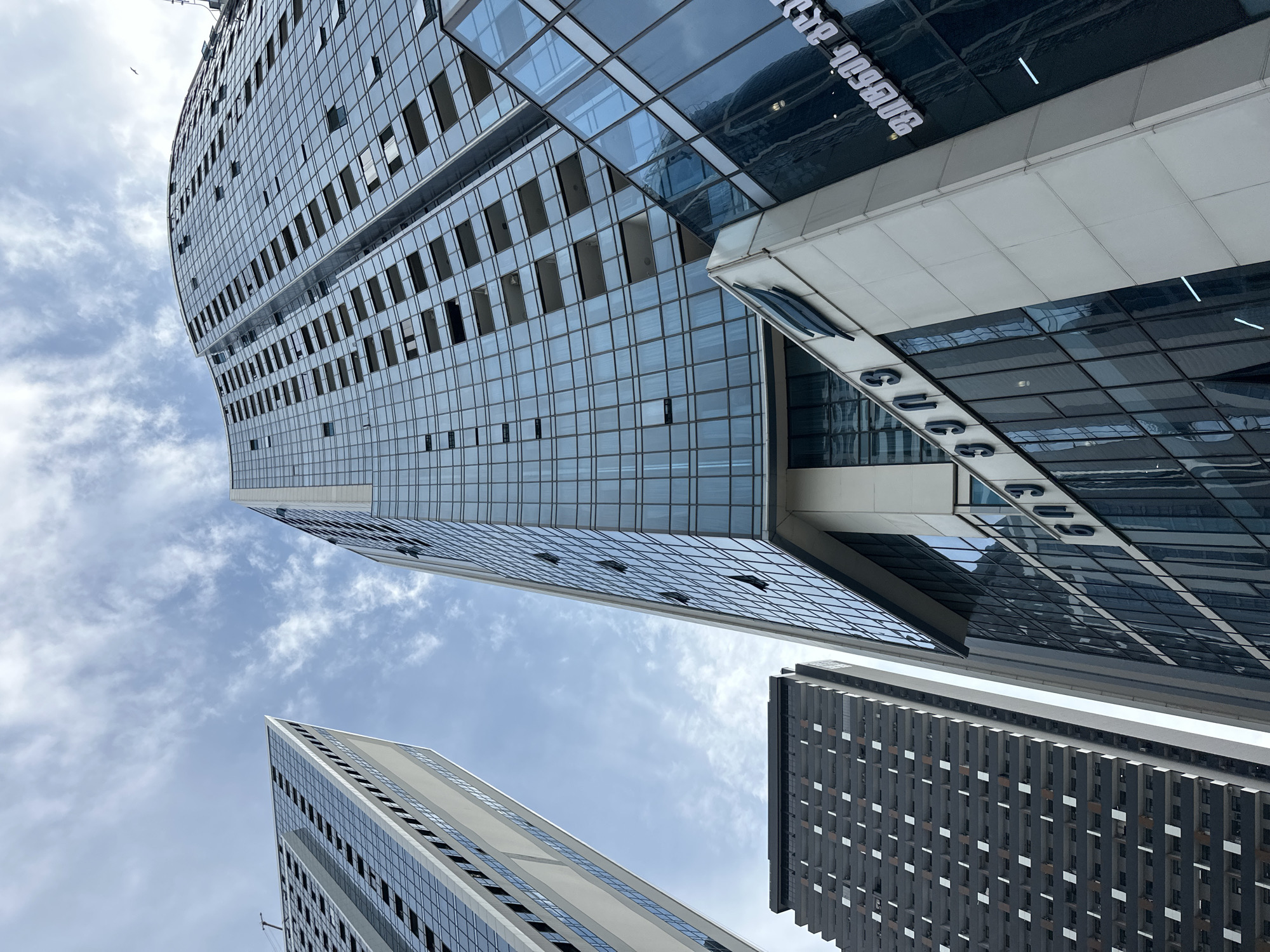 This is a low-angle, full shot of several modern high-rise buildings in Batumi, Georgia, under a partly cloudy sky. The buildings are predominantly glass and steel, reflecting the sky and surrounding structures. The building in the foreground, on the right, is a curved glass skyscraper with many open windows. Below it, a section of a white building features a sign with Georgian script and a blue logo. To the left of this foreground building, another glass skyscraper is visible, its facade made up of rectangular panes. Further to the left and in the background, a dark gray high-rise building with a grid-like pattern of balconies and white rectangular accents stands tall. The perspective emphasizes the verticality and scale of the urban architecture.