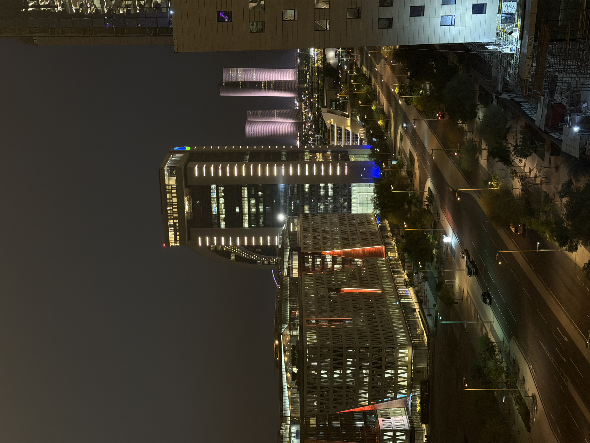 An aerial night view of a modern cityscape features several illuminated buildings and a street with traffic. The foreground is dominated by a multi-story building with a distinctive patterned facade and orange vertical lighting accents. To the left, a curved building with vertical white lights is visible. In the mid-ground, a street runs parallel to these structures, with cars and streetlights lining its path. Beyond the street, other buildings are visible, including a cluster of three cylindrical towers that are lit with a pinkish hue and appear to have a textured facade. A tall building, partially obscured on the right side of the frame, exhibits a glass exterior with reflections of city lights. Construction scaffolding is visible at the bottom right of the image. The sky is dark, indicating nighttime, and there is a slight haze.