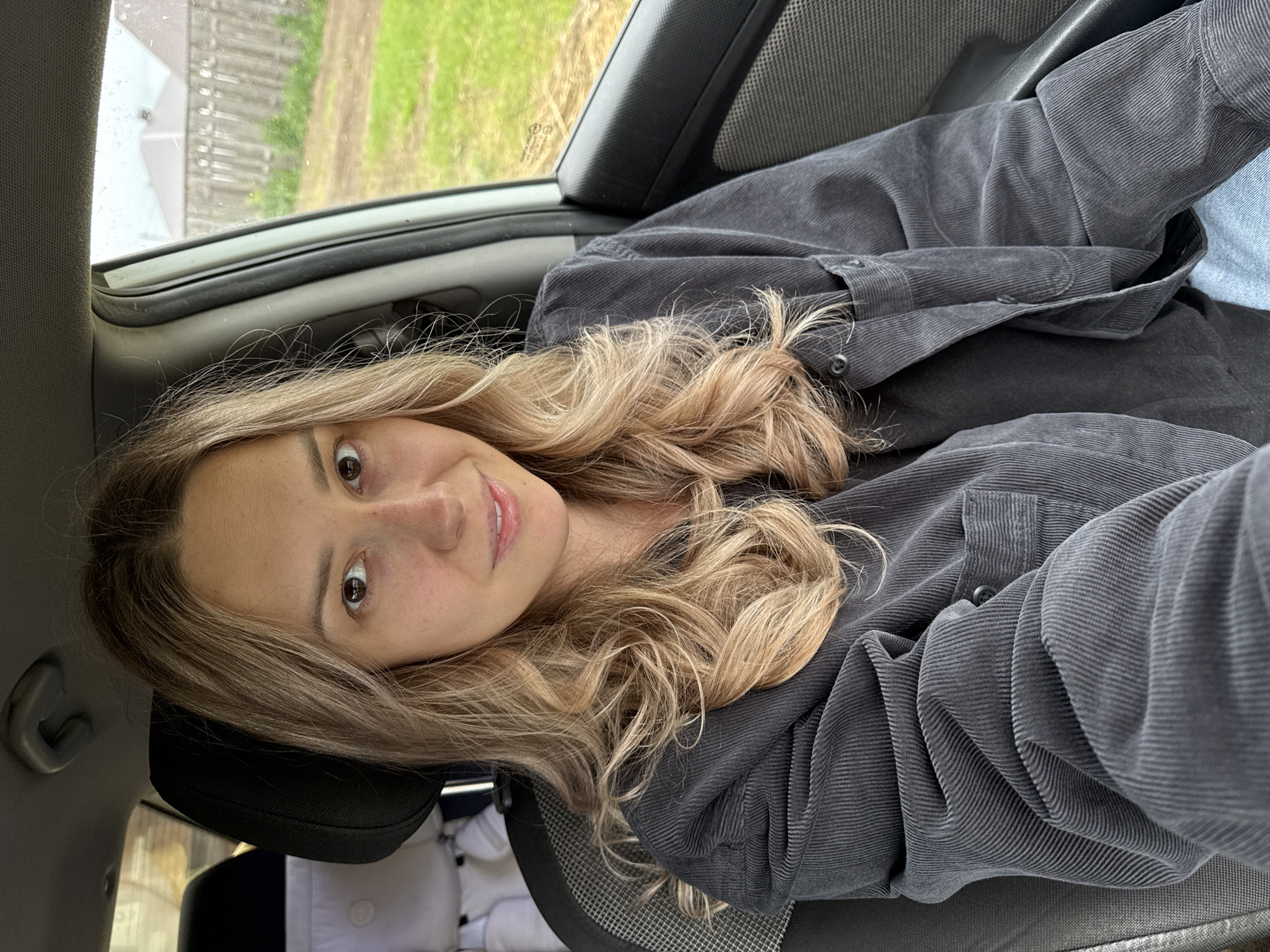 A young woman is captured in a selfie taken inside a car, looking directly at the camera with a gentle, pleasant smile. She has long, wavy blonde and light brown hair and fair skin. She is wearing a dark grey corduroy button-up shirt or jacket, with a glimpse of a light blue garment visible to her left.
The setting is the interior of a car, with the dark grey seat fabric and headliner partially visible. Through the car window, an outdoor scene unfolds, featuring green grassy areas, possibly a dirt path, and blurred foliage or distant structures. The window appears wet, with raindrops or streaks of water, indicating that it is either raining or has recently rained. The lighting suggests it is daytime, likely overcast given the soft, diffused light. The image is rotated, showing the woman leaning back or lying down. No visible text is present. This scene is located in Chaikovsky, Russia.