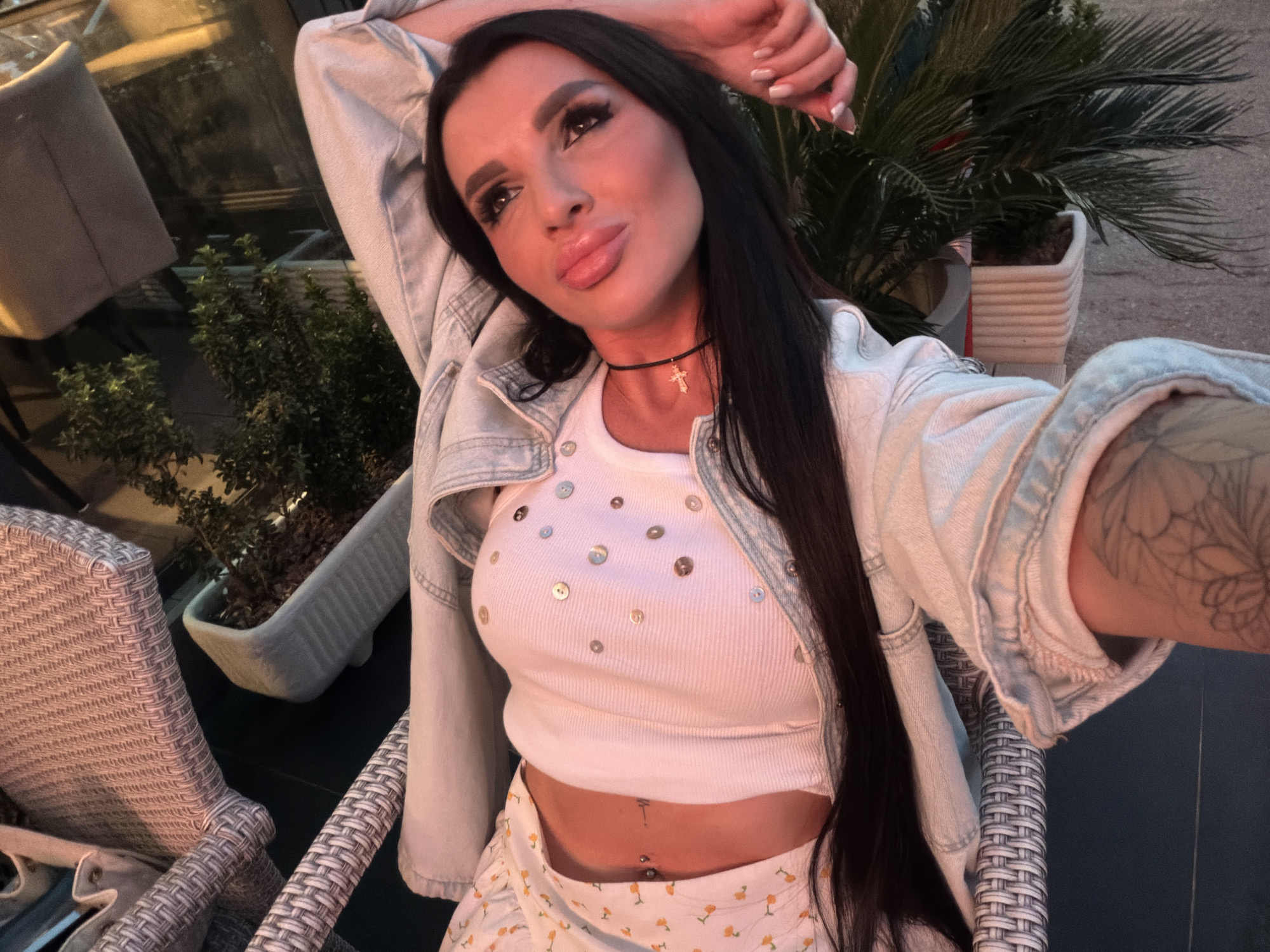 A woman is seated in a woven chair on what appears to be a patio or outdoor seating area, possibly in Budva, Montenegro. She is wearing a white cropped top with decorative buttons, a light-wash denim jacket, and a patterned skirt, exposing her midriff with a visible navel piercing. She also wears a black choker with a cross pendant. Her dark hair is long and straight. She has her arms raised, with one hand behind her head, and her face is tilted as if taking a selfie. Tattoos are visible on her arm. In the background, there are plants in pots, suggesting an urban setting with outdoor amenities. The lighting suggests it may be evening.
