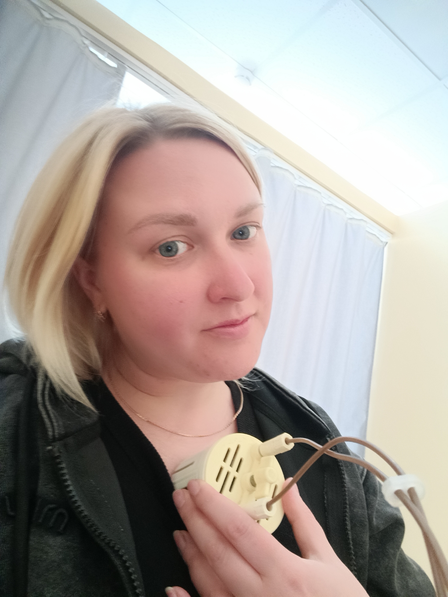 A fair-skinned woman with blonde hair is holding a beige electronic device against her chest. She is wearing a dark zip-up jacket and a gold necklace. Behind her are white curtains and a light-colored wall. The location is unknown, and there is no indication of it being in Chaikovsky, Russia.
