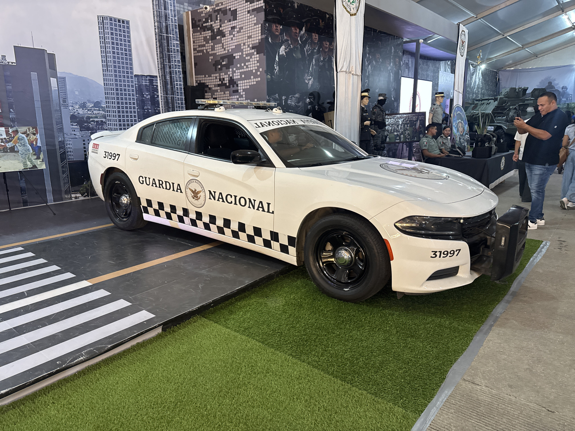 A white Dodge Charger is displayed in a tent at what appears to be an exhibition. The car is painted in the colors and markings of the Mexican National Guard (Guardia Nacional). It has the number "31997" painted on both the front and rear fenders. A black and white checkered stripe runs along the base of the doors. The car is on a display surface with a pedestrian crosswalk pattern and a patch of artificial turf. In the background are several displays, including what appear to be military mannequins, a table with uniformed figures, and displays featuring military equipment and photographs. Several people are observing the exhibit, with one person taking a photo of the car. A partial urban backdrop is visible behind the car, suggesting it may be set up to mimic a city environment. Based on the Guardia Nacional vehicle and general aesthetics, the location may be Puebla, Mexico.