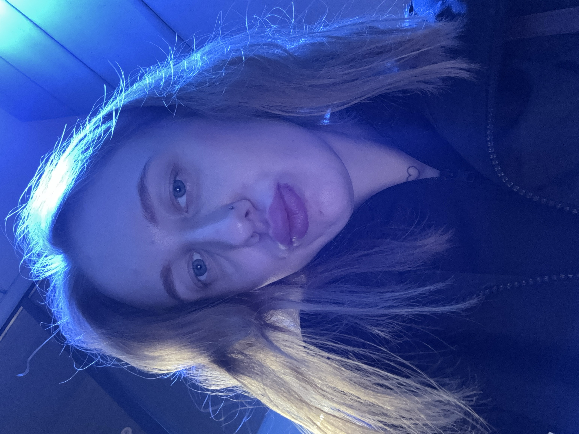 A close-up, rotated image shows a young woman with fair skin and light blonde hair. The lighting is primarily blue, casting a cool tone over the scene. She has blue eyes, a nose piercing on one side, and her lips are slightly parted. A small heart tattoo is visible on her neck. She appears to be wearing a dark jacket or top with a visible zipper and she is indoors. There is no information available about the location, or city Ol'ginskaya, Russia.