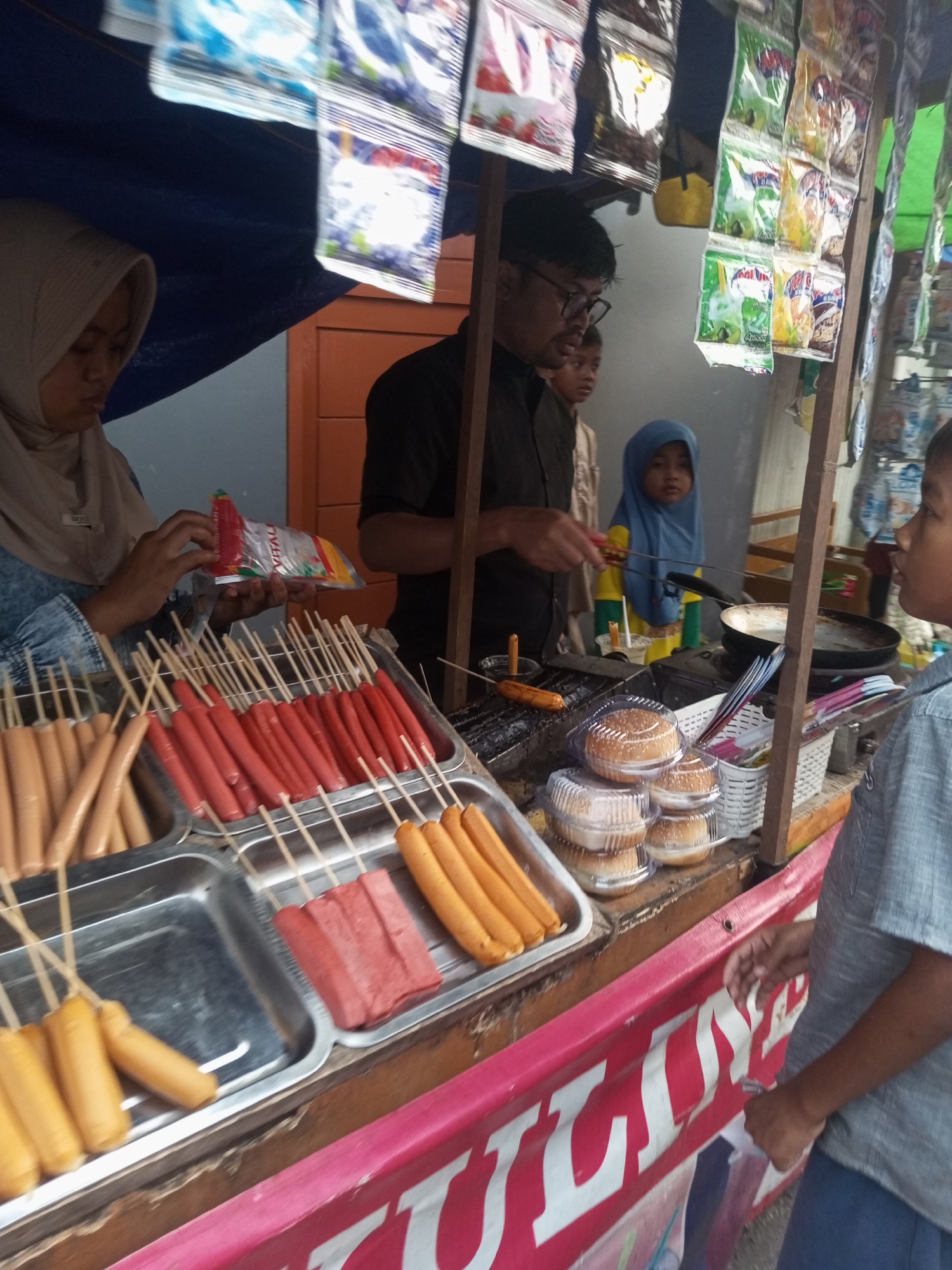 This image depicts an outdoor food stall in Pandansari, Indonesia. The stall is selling various food items, primarily sausages and burgers. In the foreground, there are several trays filled with different types of sausages on skewers. To the right, burgers are displayed in plastic containers.
Several people are present at the stall. A woman wearing a hijab is seen holding a package of what appears to be seasoning or sauce. A man, possibly the vendor, is interacting with the food. Two children are also visible; one boy is looking towards the food, and a younger child in a blue hijab is standing near the cooking area.
The stall is covered by a blue tarp, and various snack packages are displayed hanging above the food. A cooking station with a pan is set up behind the counter. The environment appears to be a bustling street scene, with the food stall set up along a roadside. The overall impression is one of a local food vendor operating in a community setting.