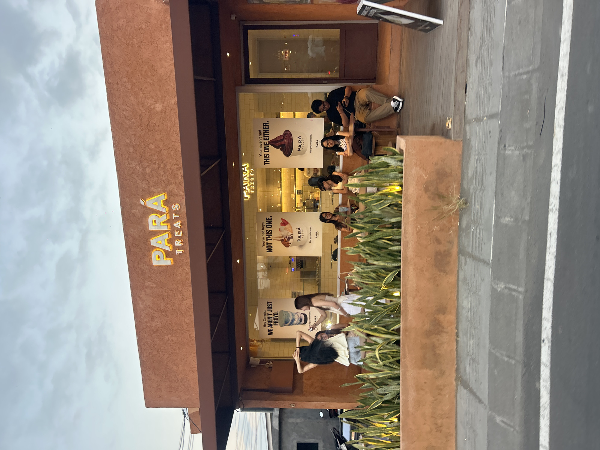 The image shows a storefront for "PARA TREATS" in North Kuta, Indonesia. The building has an orange-brown textured facade with a dark brown awning. Above the entrance, the "PARA TREATS" logo is illuminated in yellow.
The storefront features large glass windows displaying various advertisements. On the left, a poster reads "Hey Canggu, WE AREN'T JUST FROY0." to the right of it is a poster with a drink and text that says "Not your standard." and "PARA". Further to the right, another poster shows a swirl of dark ice cream and the text "You haven't had THIS ONE EITHER." accompanied by "PARA TREATS" and "Not your standard.".
Inside the store, a counter and shelving are visible. Several people are seated outside the store on a wooden deck. A woman with long dark hair is standing on the left, facing away from the camera and adjusting her hair. Two women are standing near the window, one in a white top and the other in a light yellow top. Three women are seated at a table on the left side of the deck, and one man is seated at a table on the right, looking at his phone.
In front of the storefront, there is a planter box filled with green snake plants. The area in front of the planter is paved, with a road visible in the foreground. The sky above is overcast.