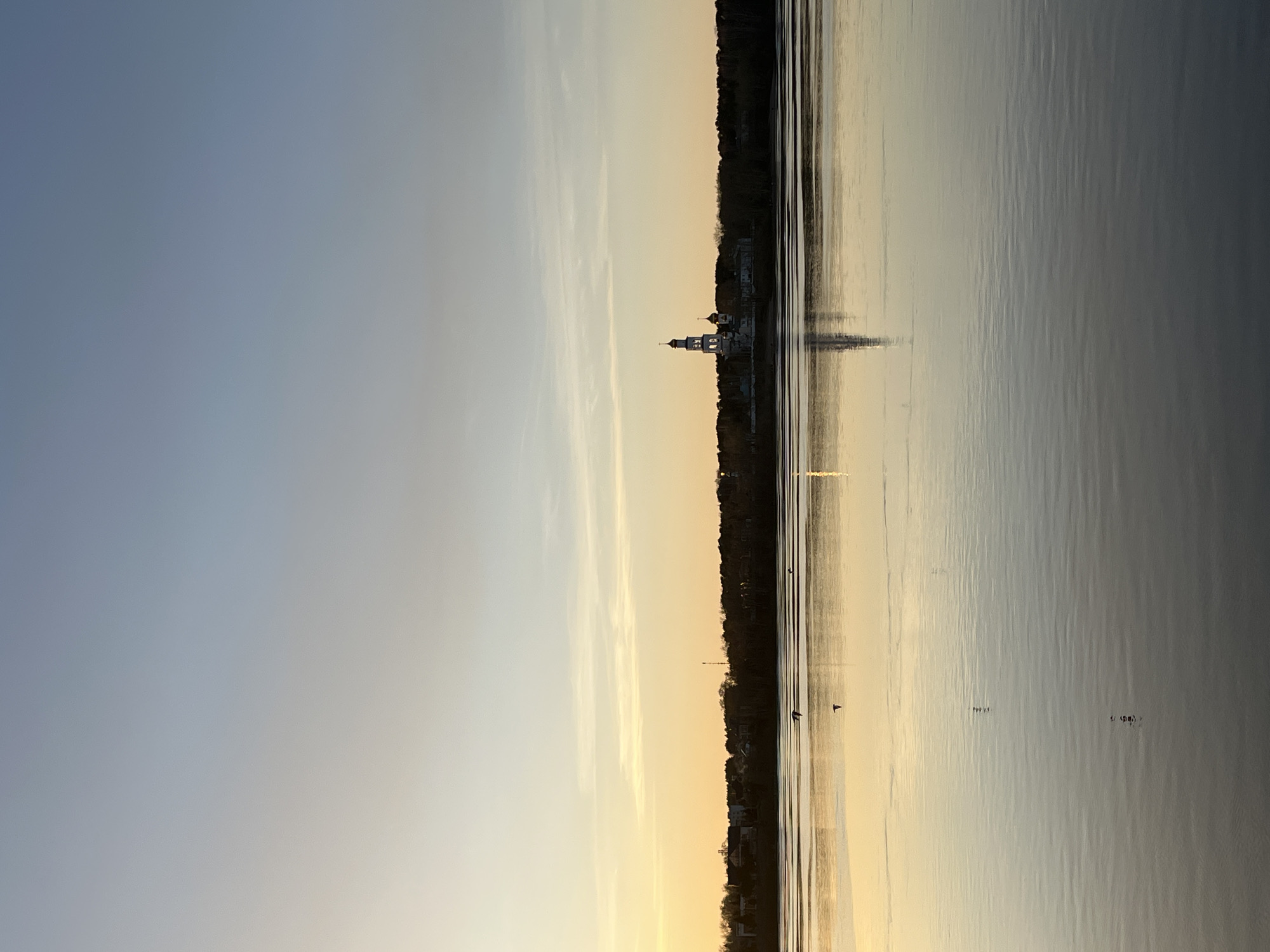 The image depicts a tranquil scene in Perm, Russia, at sunset, along the Kama River. A historical church is visible on the river bank amidst trees. The sky transitions from light blue to a warm golden hue near the horizon, reflected in the calm river water. A few birds are visible on the water surface.