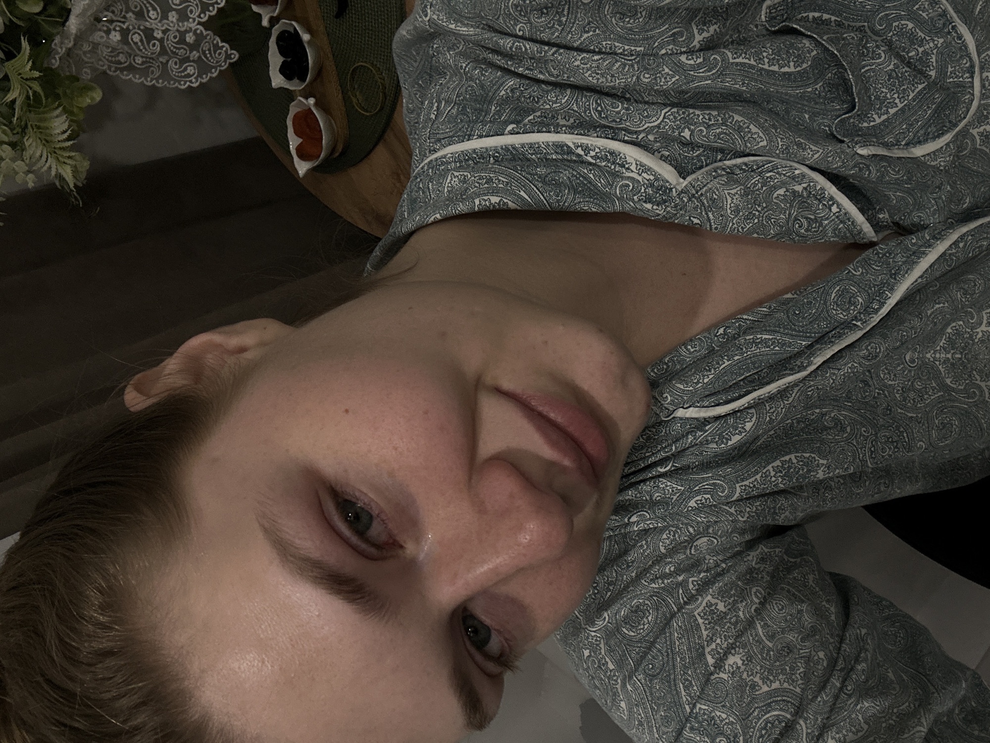 One light-skinned female individual is depicted in a close-up, angled portrait. Her light brown hair is pulled back. Her eyes are green-blue, displaying redness around the conjunctiva, with a single tear visible in the medial canthus of her left eye. Her facial expression shows a slight upturn of the lips on the right side, indicative of a partial smile. Her skin appears flushed. She is wearing a green-blue paisley-patterned pajama top with white piping along the collar and front opening. The background shows an indoor setting. A wooden surface to the upper right holds a dark green circular placemat. On the placemat are two small, white, boat-shaped ceramic bowls: one contains orange-colored dried fruit, and the other contains dark-colored dried fruit. A white lace fabric and a partial floral arrangement with green foliage and white elements are visible behind the bowls. A thin, light-colored elastic band rests on the placemat. Dark fabric, likely curtains, is present in the upper left background. The lighting is subdued. The scene is located in Kazan, Russia.