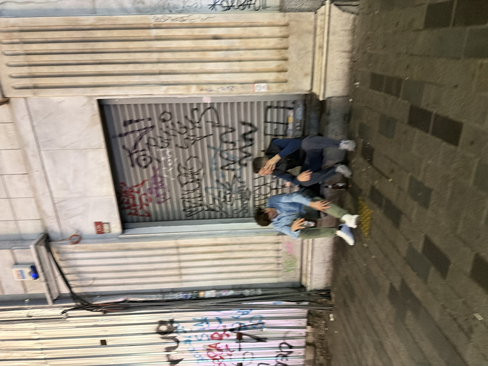 Two individuals are located on a sidewalk next to a building with a metal roll-up door covered in graffiti. One person, dressed in a dark jacket and dark pants, is positioned closer to the roll-up door. Another individual, dressed in a blue denim shirt and olive green pants, is positioned adjacent. The building also has a corrugated metal awning above the roll-up door. The sidewalk is made of rectangular stone blocks. The scene is consistent with an urban environment, potentially in Beyoğlu, Turkey, based on architectural elements and building style.