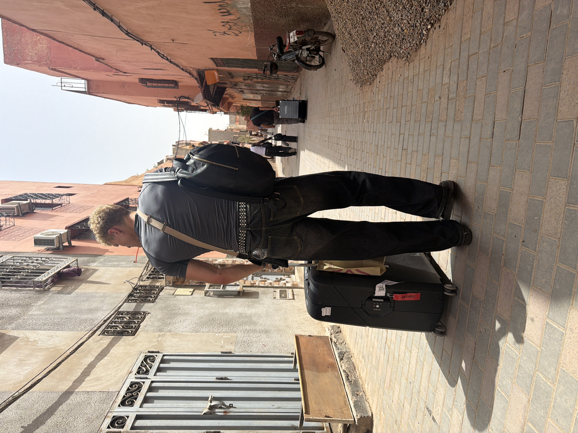 The image depicts a narrow, sunlit street in Marrakesh, Morocco, with buildings made of reddish-pink and beige stucco lining both sides. The street is paved with tan and gray brick. In the foreground, a person with blond, curly hair is walking away from the camera, pulling a black rolling suitcase. They are wearing a dark gray t-shirt, dark wash jeans, and a studded belt. A black backpack is slung over their left shoulder, and a beige strap of another bag is visible. Two paper shopping bags hang from the suitcase handle. In the middle ground, two other figures are walking further down the street, one pulling a suitcase and the other carrying a bag. A small, dark-colored motorcycle is parked on the right side of the street, near a pile of gravel. The architecture features wrought-iron window grates and a visible air conditioning unit on one of the buildings. The overall impression is of a typical, warm day in a Moroccan medina.