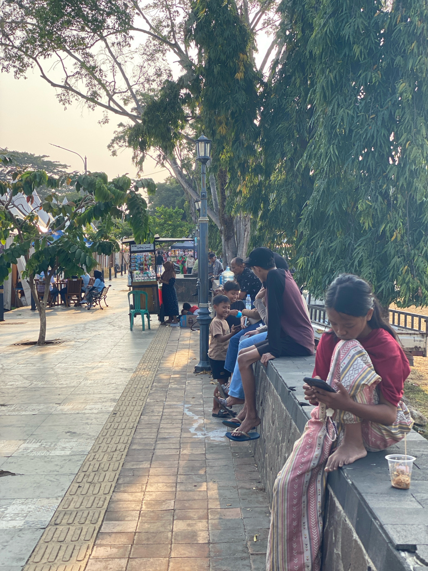 The image depicts a street scene in the city of Nyamok, Indonesia, characterized by warm, diffused sunlight. In the foreground, a young girl sits on a low stone wall, engrossed in her mobile phone. She wears a red top and loose-fitting pants with a patterned design. A plastic cup containing a small amount of food sits next to her. Behind her, several other individuals are seated on the same wall, some interacting with each other and others looking down. Among them are men and children. One child in a light brown t-shirt appears to be interacting with an adult.
The environment includes a paved walkway with tactile paving for the visually impaired. Further in the background, there are trees with dense foliage, providing shade. A street lamp with a decorative design is visible, as well as several vendor stalls. Some people are seated at tables in an outdoor seating area to the left, suggesting a commercial or recreational space. The overall impression is one of a public area with people casually spending time outdoors.