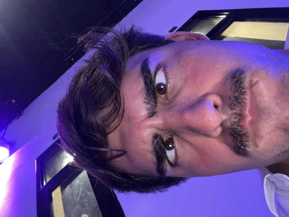 A close-up selfie shows a man with dark hair and a mustache. He looks directly at the camera with wide eyes and slightly pursed lips, seemingly making a funny face. His skin is flushed, possibly due to lighting. The picture is taken at an unusual angle, tilted significantly, giving a unique perspective. A hint of white clothing is visible at the bottom corner. A window frame and a light source are in the background, illuminated by blue and white light.
