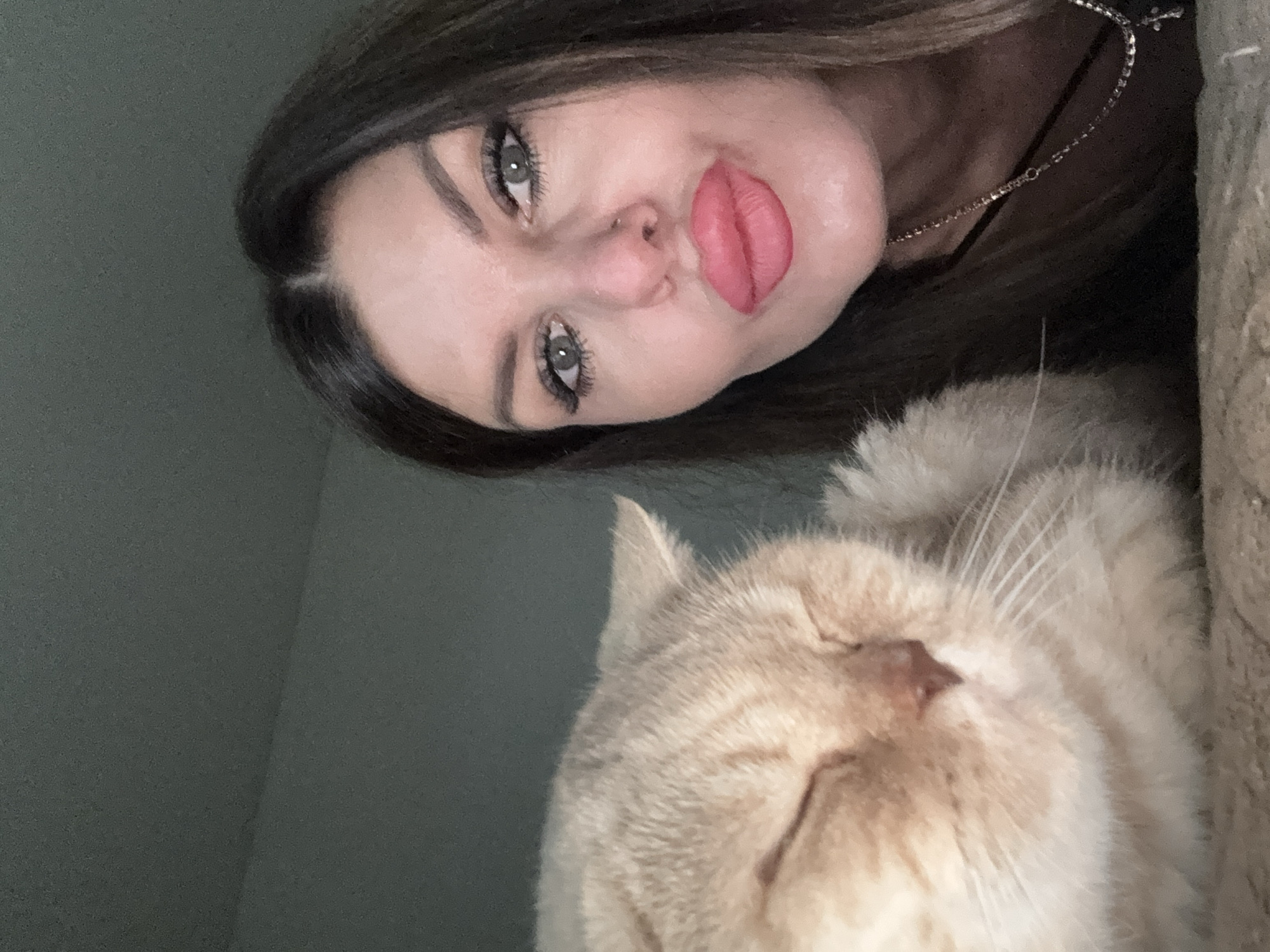 A selfie features a woman with dark hair, green eyes, and bright pink lipstick, puckering her lips. She is positioned to the right of a light brown cat with its eyes closed, positioned on the left. The background is a plain, dark gray wall, indicating an indoor setting. The image was captured in Chaikovsky, Russia.