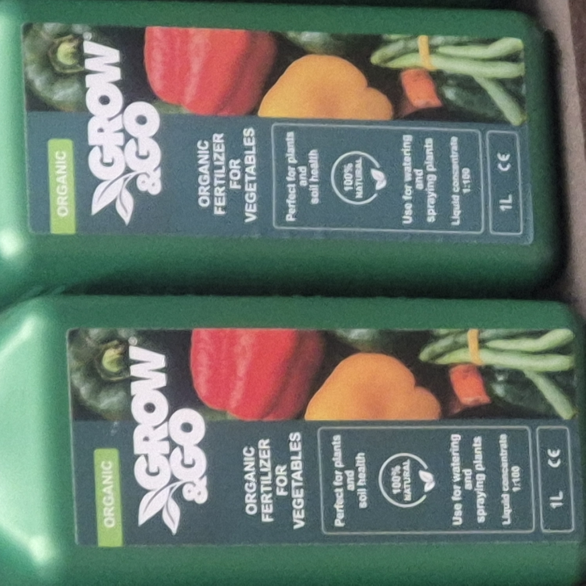 This media file contains two green plastic bottles of "GROW & GO" organic fertilizer for vegetables. The labels on the bottles feature images of red and yellow bell peppers, and green beans. Text on the labels indicates the product is organic, perfect for plants and soil health, suitable for watering and spraying, and is a liquid concentrate with a dilution ratio of 1:100. The bottles are 1L in volume and have the CE mark. The bottles are presented in a top-down view, with one bottle slightly overlapping the other. The setting appears to be indoors, possibly a storage area or retail display, given the lighting and the rough texture of the surface beneath the bottles. There is no discernible time of day, weather, or emotional context visible. The location context provided is Nowa Wieś Książęca, Poland.