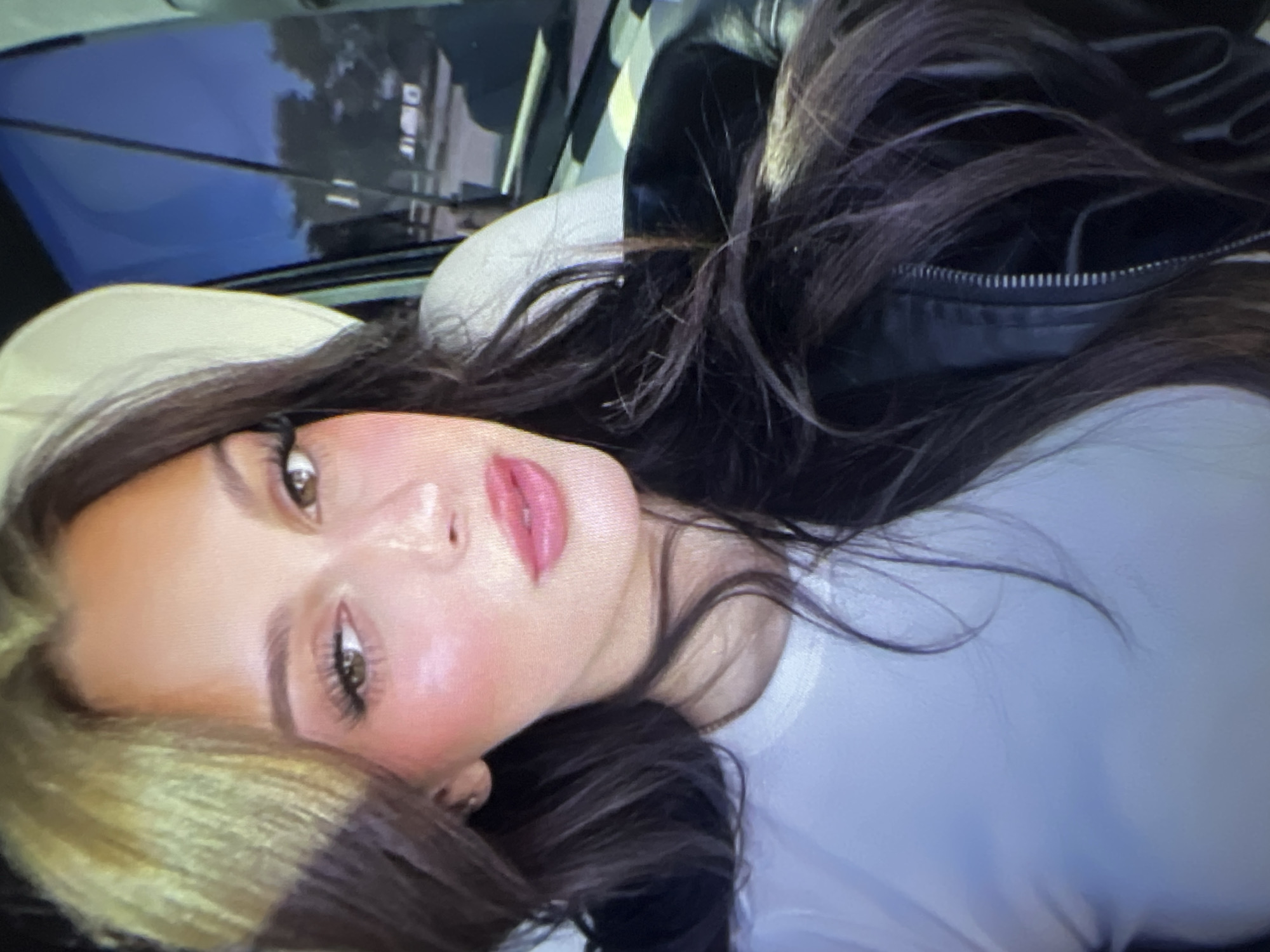 A woman with long, dark brown hair, some of which is dyed blonde, is looking at the camera. She is wearing a white t-shirt and a black leather jacket. She has on makeup, including false eyelashes and bright pink lipstick. The background appears to be the interior of a car, with a seat visible on the left and a window on the right. Through the window, trees and buildings can be seen, suggesting an outdoor environment, possibly in a city. There is no specific indication of the location being Pyatigorsk, Russia.