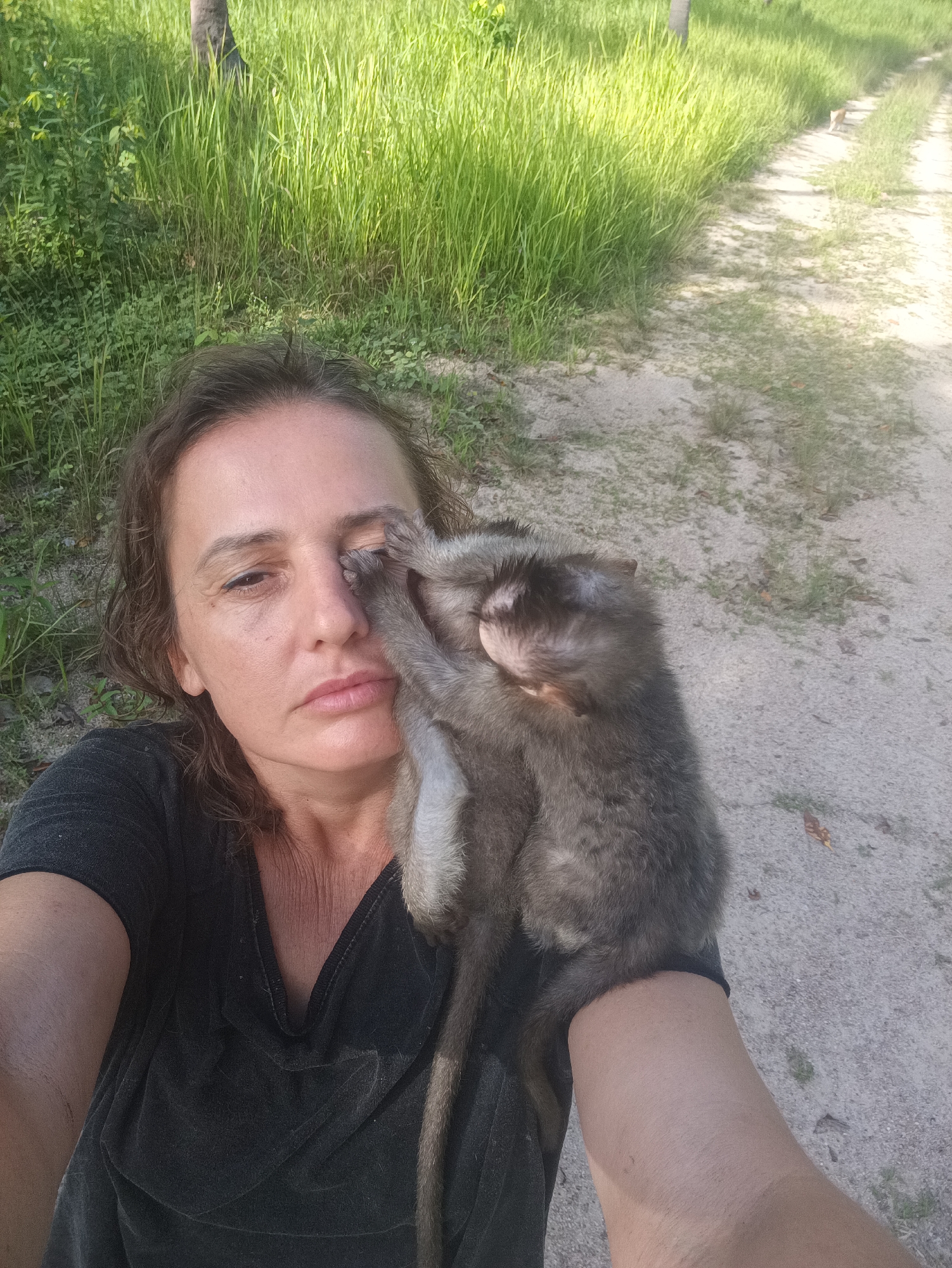 A medium shot depicts a woman holding a monkey. The woman, facing forward, has light skin, dark hair, and is wearing a dark t-shirt. The monkey, grayish-brown in color, clings to her arm and face, with its hand near her eye. The setting appears to be outdoors, with tall green grass in the background on the left side and a dirt path on the right side of the frame. The location is Baan Thong Nang, Thailand. The action is a selfie being taken by the woman with the monkey clinging to her.