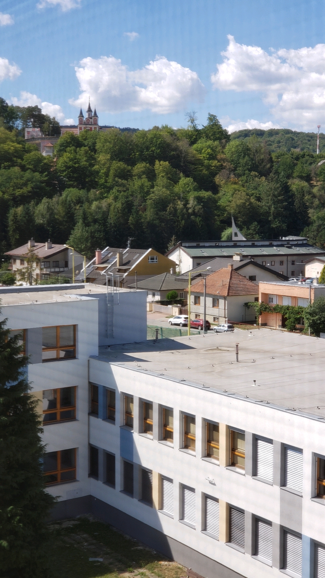This image captures a daytime view of a suburban area in Prešov, Slovakia. In the foreground, a white, multi-story building with numerous windows is prominent. The building features a flat roof with visible vents and a ladder on its side. To the left of this building, a dark green coniferous tree partially obscures the view.
Behind the foreground building, a residential area with several houses and other structures is visible. These houses have pitched roofs of various colors, including brown, yellow, and gray. Some houses have visible chimneys, and there are parked cars on the street.
Further in the background, a dense forest covers a hillside. At the top of the hill, a distinctive red and white building with multiple spires, resembling a church or monastery, is visible against a bright blue sky dotted with white clouds. A telecommunications tower is also present on a distant hill to the right. The overall impression is a tranquil, semi-rural landscape within the city of Prešov.