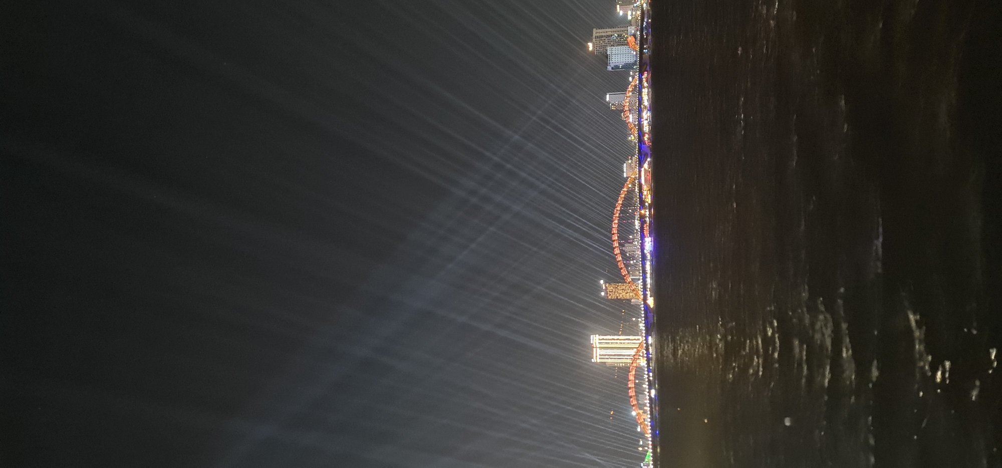 An eye-level nighttime long exposure captures the skyline and the Dragon Bridge in the city of Đà Nẵng, Vietnam, reflecting over water. The Dragon Bridge, illuminated in red and orange, arches across the water. Buildings of various heights are visible on the shore, illuminated with white and colored lights. Light beams emanate upwards from various points, forming a fan-like pattern across the sky. The water surface reflects the city lights.