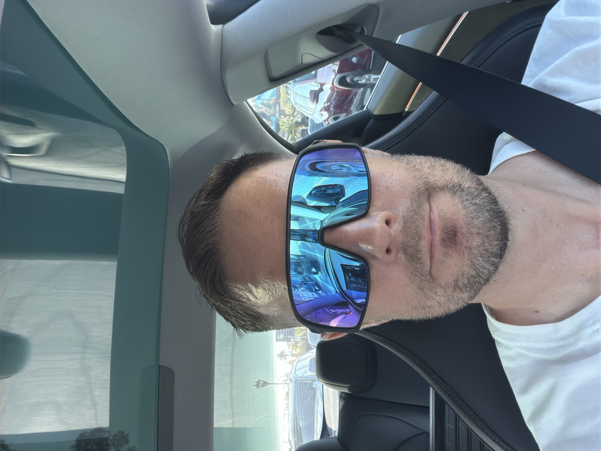 A man is sitting in the driver's seat of a car, wearing large, futuristic sunglasses. The sunglasses reflect the interior of the car and the road ahead, showing traffic and palm trees. The car has a panoramic sunroof, and the man is wearing a white t-shirt. The scene appears to be in a city, possibly Los Angeles, given the presence of palm trees and the style of the cars.