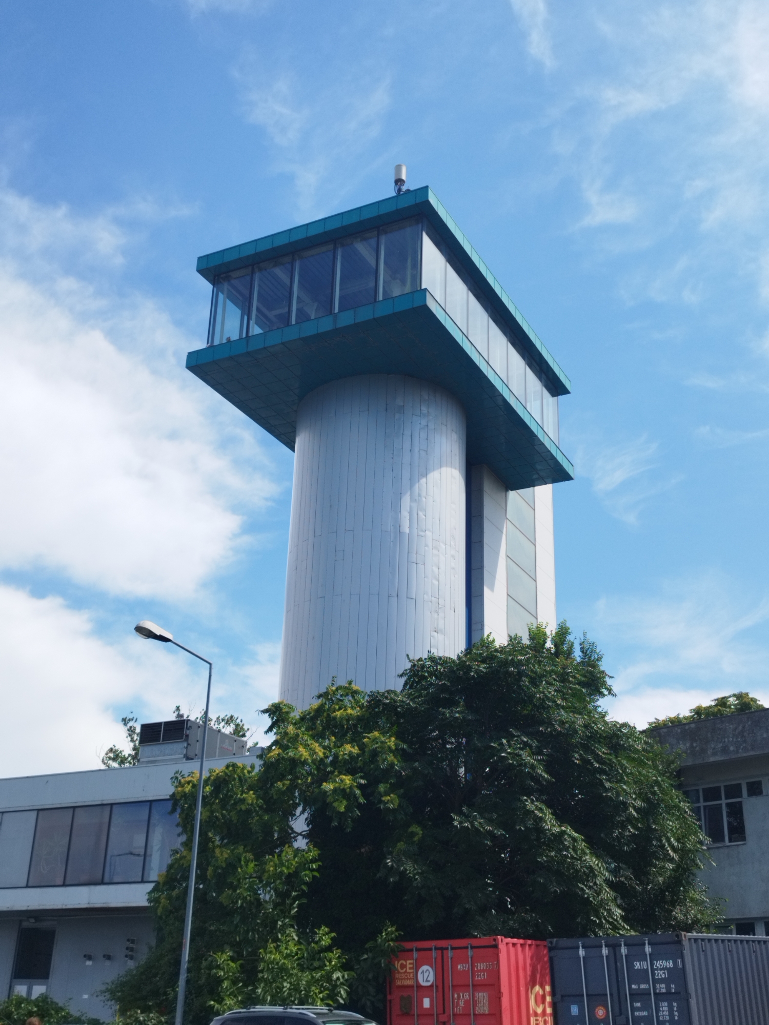 This is a low-angle, medium shot of a modern, cylindrical building with a glass-enclosed observation deck on top, under a blue sky with scattered clouds. The building is located in Constanța, Romania, specifically at the Constanța Port Control Tower.
The main structure is a tall, silver cylinder, likely made of metal, with vertical seams. The upper section is a rectangular, cantilevered platform with large glass windows on all sides, giving it a modern, almost futuristic appearance. The observation deck is painted teal blue, contrasting with the silver of the cylinder. A small, white antenna or equipment is visible on the roof of the observation deck.
In the foreground, a large, dense green tree with dark foliage partially obscures the lower portion of the building and extends into the bottom right of the frame. Below the tree and to the right, two shipping containers are visible, one red and one dark gray. The red container has white text and markings, including the number "12". The gray container has similar text and markings, with "SKIU 245968 22G1" prominently displayed.
To the left of the main tower, a portion of a lower, gray building is visible. This building has large windows and appears to be part of the port facilities. A gray street lamp with a white light fixture is positioned in front of the cylindrical tower and to the left of the tree. The sky is a clear, bright blue with wispy white clouds, indicating it's a daytime shot. The overall impression is one of industrial architecture combined with natural elements.