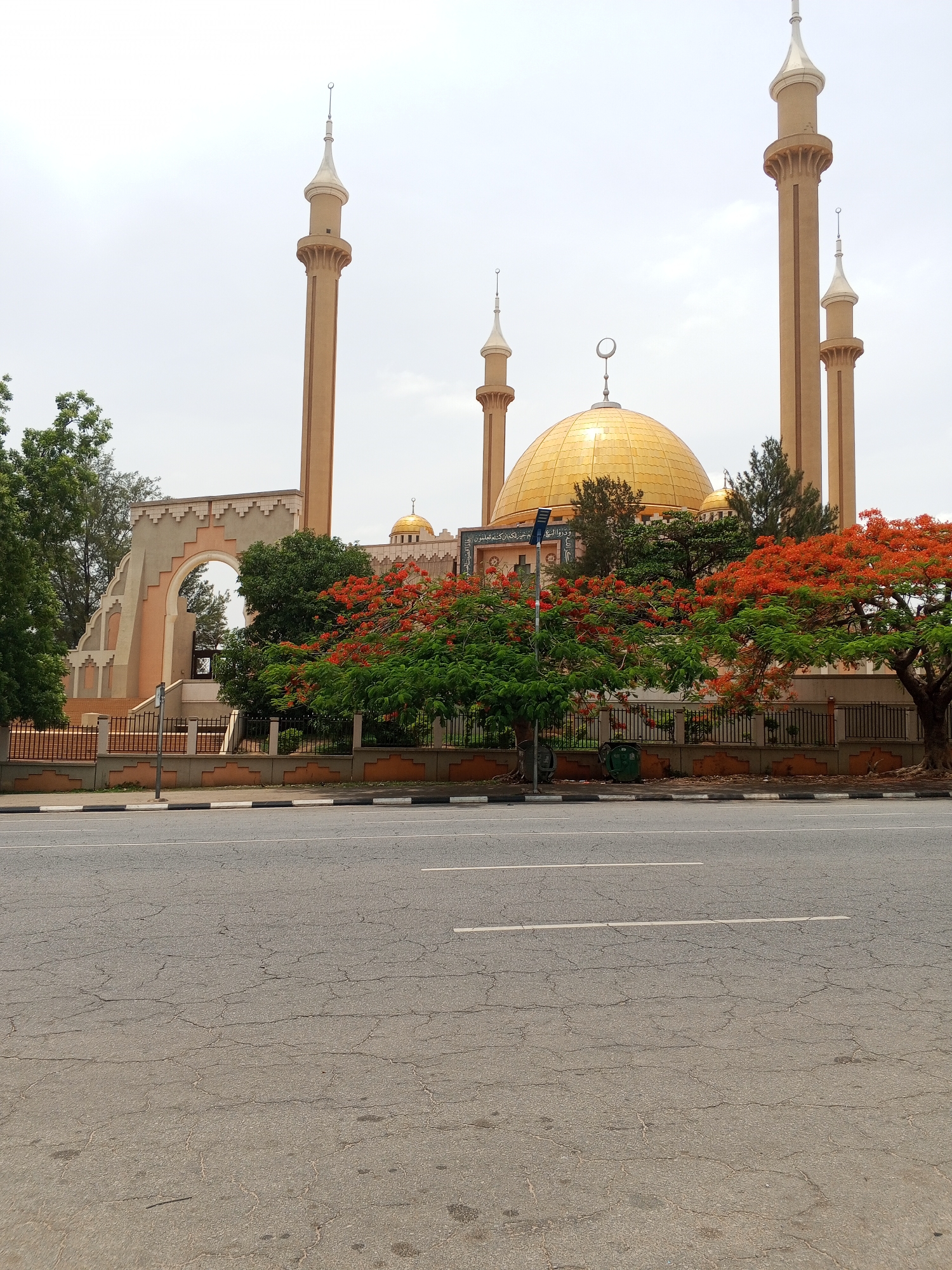 The image shows the National Mosque in Abuja, Nigeria. The mosque features a large golden dome and several tall minarets with silver tops. It is surrounded by trees, some with vibrant orange flowers. In the foreground is a road with visible cracks in the asphalt. A low fence separates the mosque grounds from the road. The sky is overcast. There are no people visible.