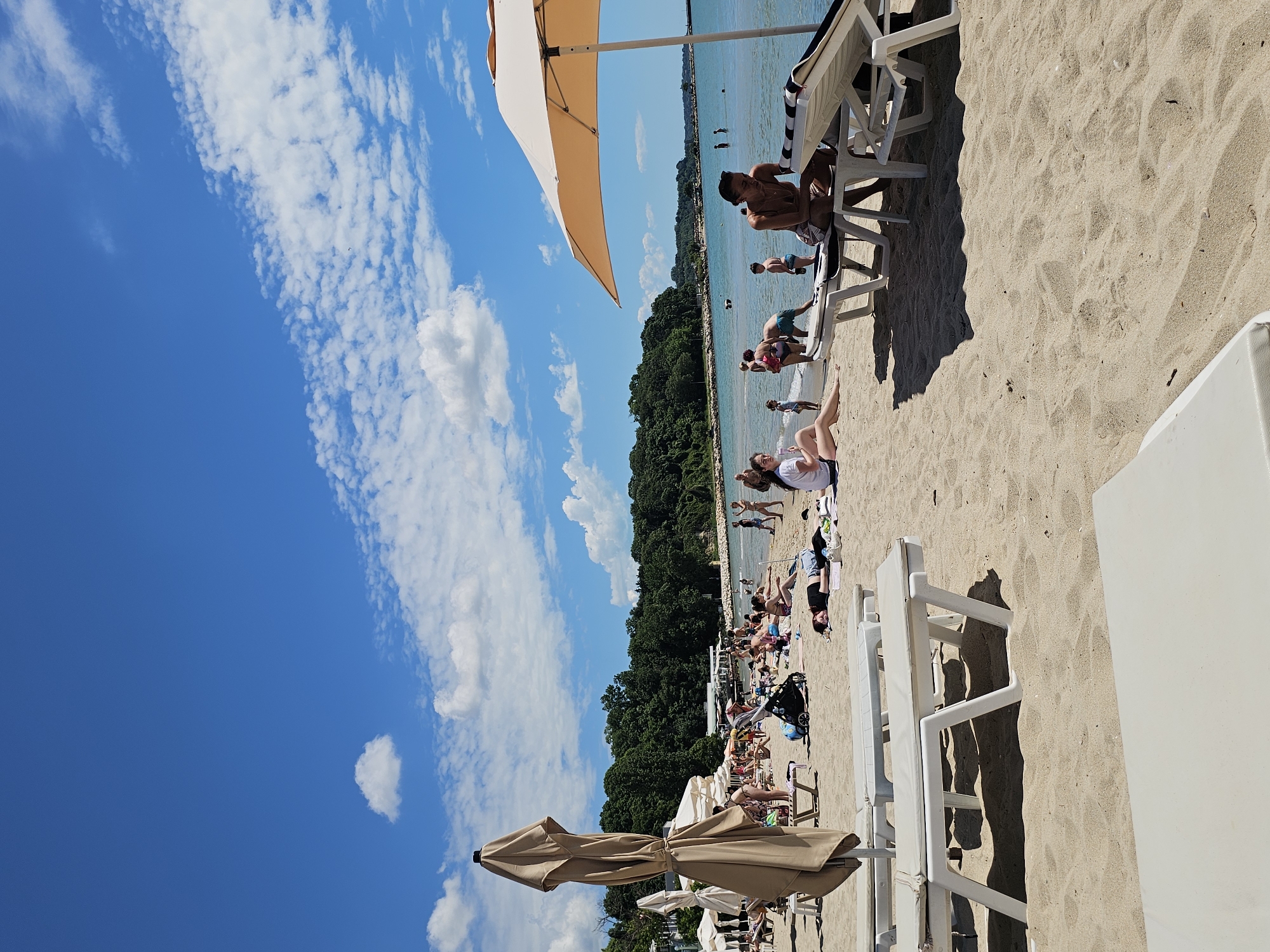 The image depicts a sunny day at a crowded beach in an unknown city. In the foreground, several white lounge chairs are arranged on the sand. A large, beige umbrella is partially visible on the right side of the frame, casting a shadow. Further back, more lounge chairs and umbrellas are scattered across the beach. Numerous people are engaged in various activities: sunbathing, relaxing on chairs, walking on the sand, and wading in the shallow water. A stroller is visible among the beachgoers. The background shows a line of lush green trees and a distant view of the sea under a bright blue sky with scattered white clouds. The overall scene conveys a typical summer beach day with many people enjoying the sun and water.