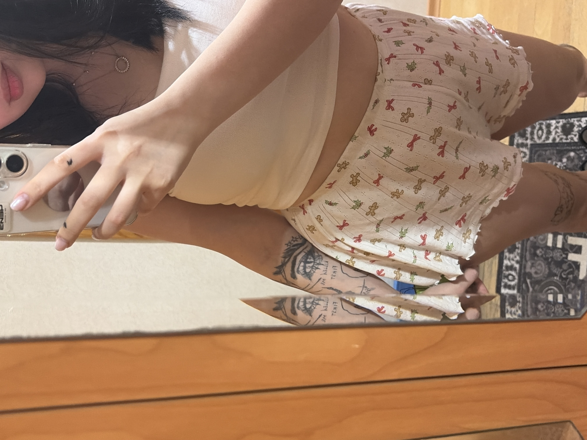 This media file captures a mirror selfie of a person, likely a young female, taken indoors in Chaikovsky, Russia. The person is visible from the chest down, with only a partial view of her face, including her lips and chin. She is wearing a white, sleeveless crop top and white shorts adorned with a festive pattern of red bows, green holly leaves, and gingerbread men, featuring scalloped edges.
She is holding a smartphone, seemingly an iPhone, in her left hand to take the selfie, with her fingers wrapped around the device. A small black heart tattoo is visible on one of her fingers, and she wears a ring on another. Around her neck is a delicate necklace with a small, circular, possibly studded, pendant.
A notable detail is a large, intricate tattoo on her upper right leg/thigh, which depicts stylized eyes with prominent eyelashes and contains partially visible text that appears to read "THTY WE TRXT." The setting appears to be a room with light-colored walls and wooden furniture, likely a dresser or table, reflecting in the mirror. A dark, patterned rug or carpet is also visible in the reflection on the floor. The image is taken at an angle, possibly due to the mirror's position or the camera's tilt. Text "N E" is faintly visible on the phone.