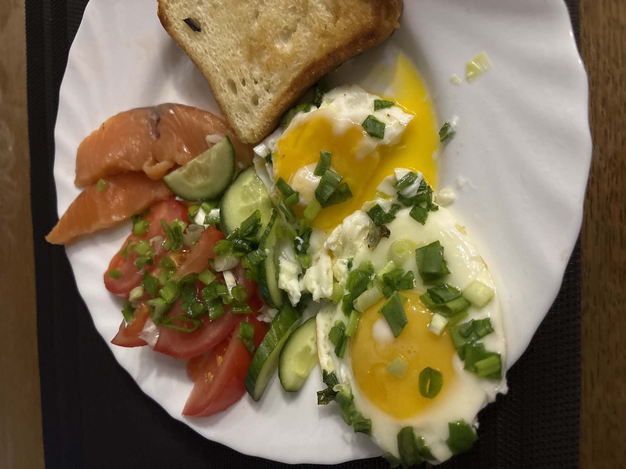 This close-up, overhead photograph showcases a freshly prepared meal served on a white, fluted plate. The meal appears to be a hearty breakfast or brunch, composed of several distinct components.
The plate features two sunny-side-up fried eggs, distinguished by their vibrant yellow yolks and crisp white edges. Both eggs are generously garnished with finely chopped green onions, with one yolk visibly broken, releasing its rich liquid onto the plate.
Adjacent to the eggs, a colorful salad composed of thinly sliced red tomatoes and green cucumbers is arranged. This salad is also sprinkled with green onions and interspersed with crumbled white, feta-like cheese. On the left side of the plate, several delicate slices of orange-pink fish, likely smoked salmon, are neatly presented. A single slice of golden-brown toasted bread completes the ensemble, positioned at the top of the plate.
The plate rests on a dark, textured surface, possibly a placemat, with a warm-toned wooden surface visible in the background, suggesting an indoor setting such as a home or café. The lighting is bright and even, highlighting the fresh ingredients and appetizing presentation of the food. There are no people visible, and no visible text in the image. The time of day is implied to be morning or early afternoon due to the nature of the meal.