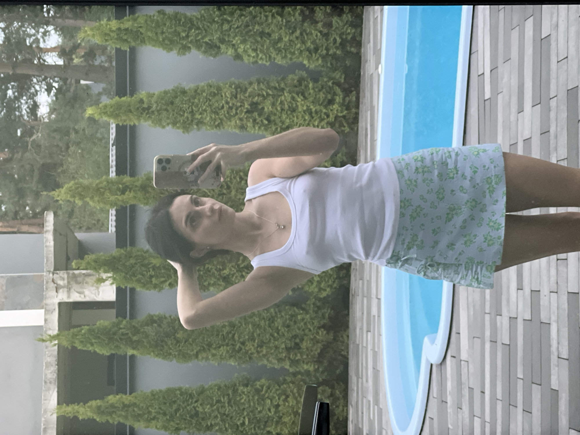 A young woman stands in front of a swimming pool, holding a smartphone in her right hand and taking a selfie. She is wearing a white tank top and a light blue skirt with a floral pattern. The woman has her left arm raised and her hand resting on her head. The swimming pool is rectangular and filled with blue water. The ground around the pool is paved with gray tiles. Behind the woman, there are several tall, green evergreen trees. In the background, there is a building with a gray exterior and a concrete structure. The scene appears to be an outdoor residential area.