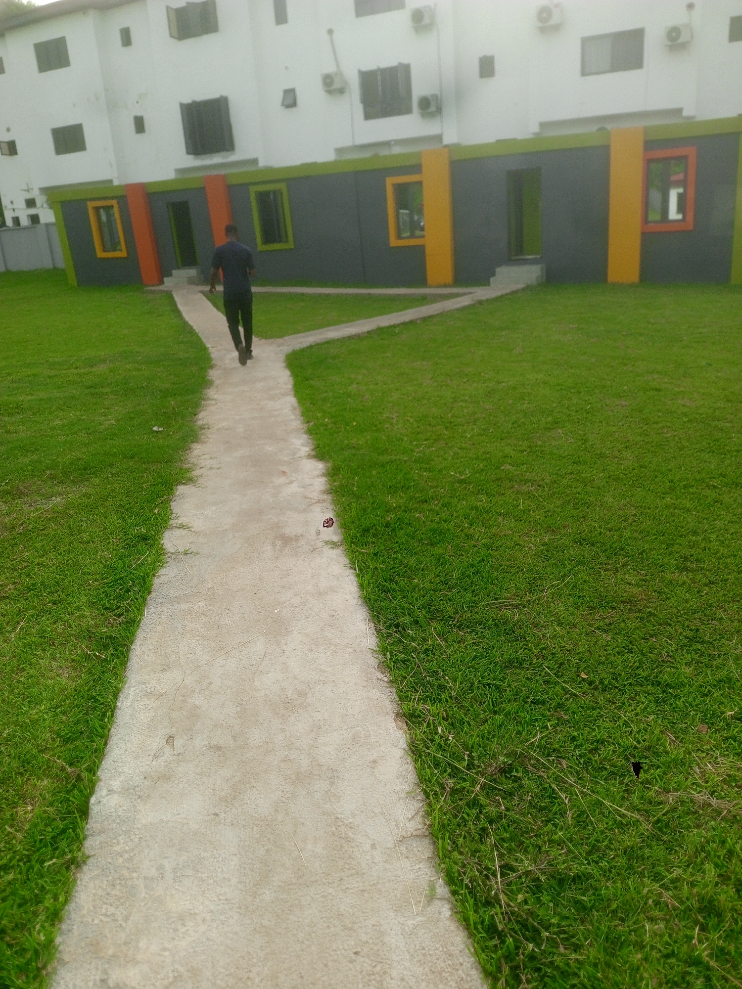 This image captures an outdoor scene in Gwarinpa, Nigeria, featuring a person walking on a concrete pathway through a grassy area towards a building complex.
The foreground and mid-ground are dominated by a lush green lawn, neatly mown. A light grey concrete pathway curves gently from the bottom center of the frame and then forks, with one branch veering to the left and another continuing straight ahead, towards the buildings. The person is seen from behind, wearing a dark blue shirt and dark trousers, walking briskly along the central path.
In the background, there are two distinct buildings. Above, a multi-story white building with numerous dark-framed windows and several visible outdoor air conditioning units forms a prominent backdrop. Below it, a single-story structure with a contemporary design stands out. This building features a dark grey wall, highlighted by bright lime green strips along its top and bottom edges. Vertical orange pillars punctuate the facade, framing sections that include rectangular windows with yellow, orange, and red frames. A dark green door is visible in the center of this lower building, aligning with the path.
The lighting suggests it is daytime, with no strong shadows, indicating either an overcast sky or midday sun. The overall scene conveys a well-maintained environment, possibly part of a modern residential or commercial complex. There is no visible text in the image.