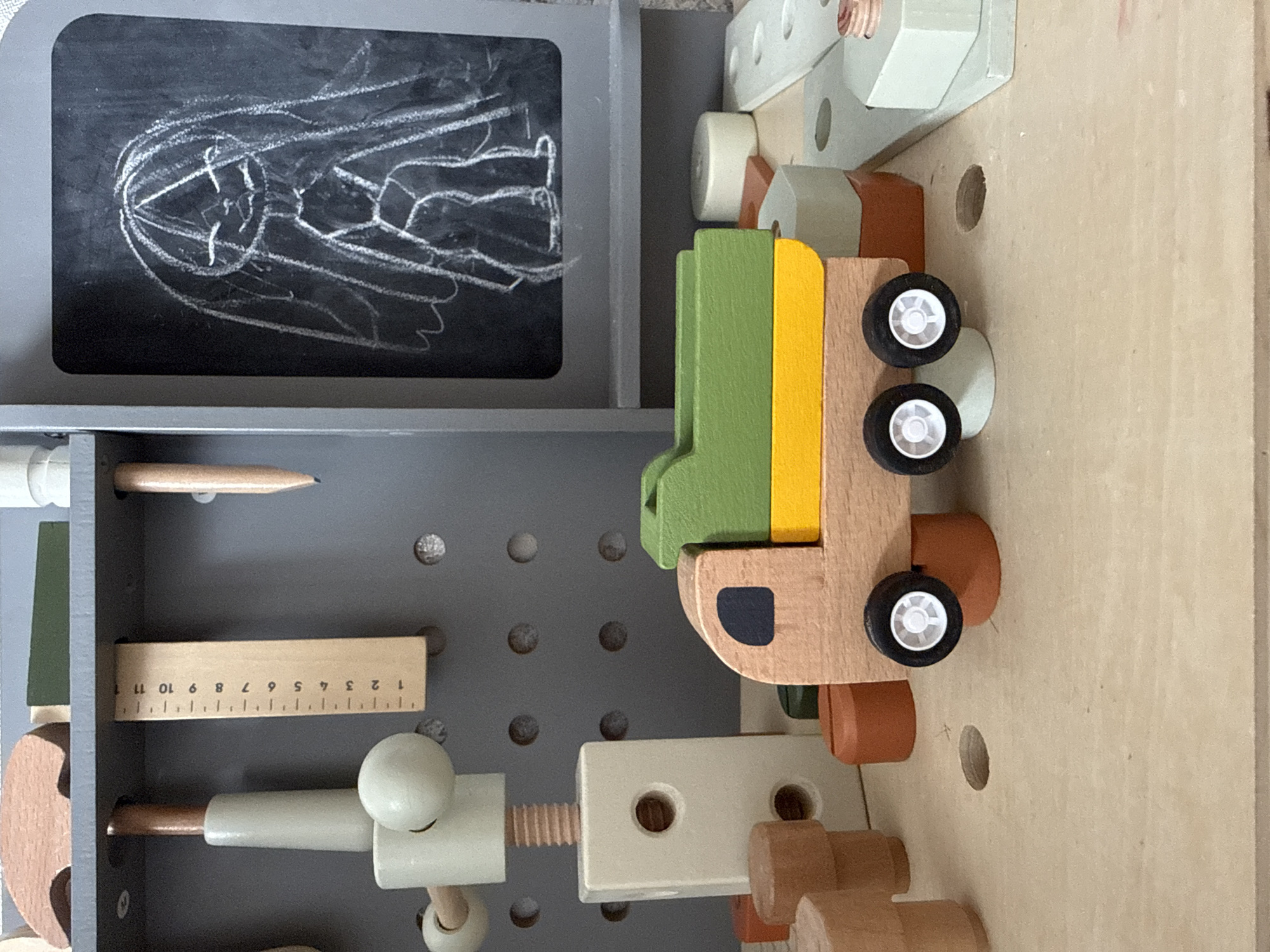 The image shows a close-up view of a wooden toy workbench. The workbench is gray with multiple round holes on its surface.
On the top left of the image, there is a small chalkboard with a white chalk drawing of an object. Below the chalkboard, there is a shelf that contains a pencil and a small wooden ruler marked with numbers 1 through 11. Further below, there is a wooden toy screw, clamp and other assorted wooden pieces.
On the right side of the image, there is a multi-colored wooden toy truck with black wheels. There are other wooden toy shapes scattered around the truck.
The background surface appears to be a plain, light-colored wooden table or surface.
There is no information that indicates the location of this scene is Rusanovo, Russia.