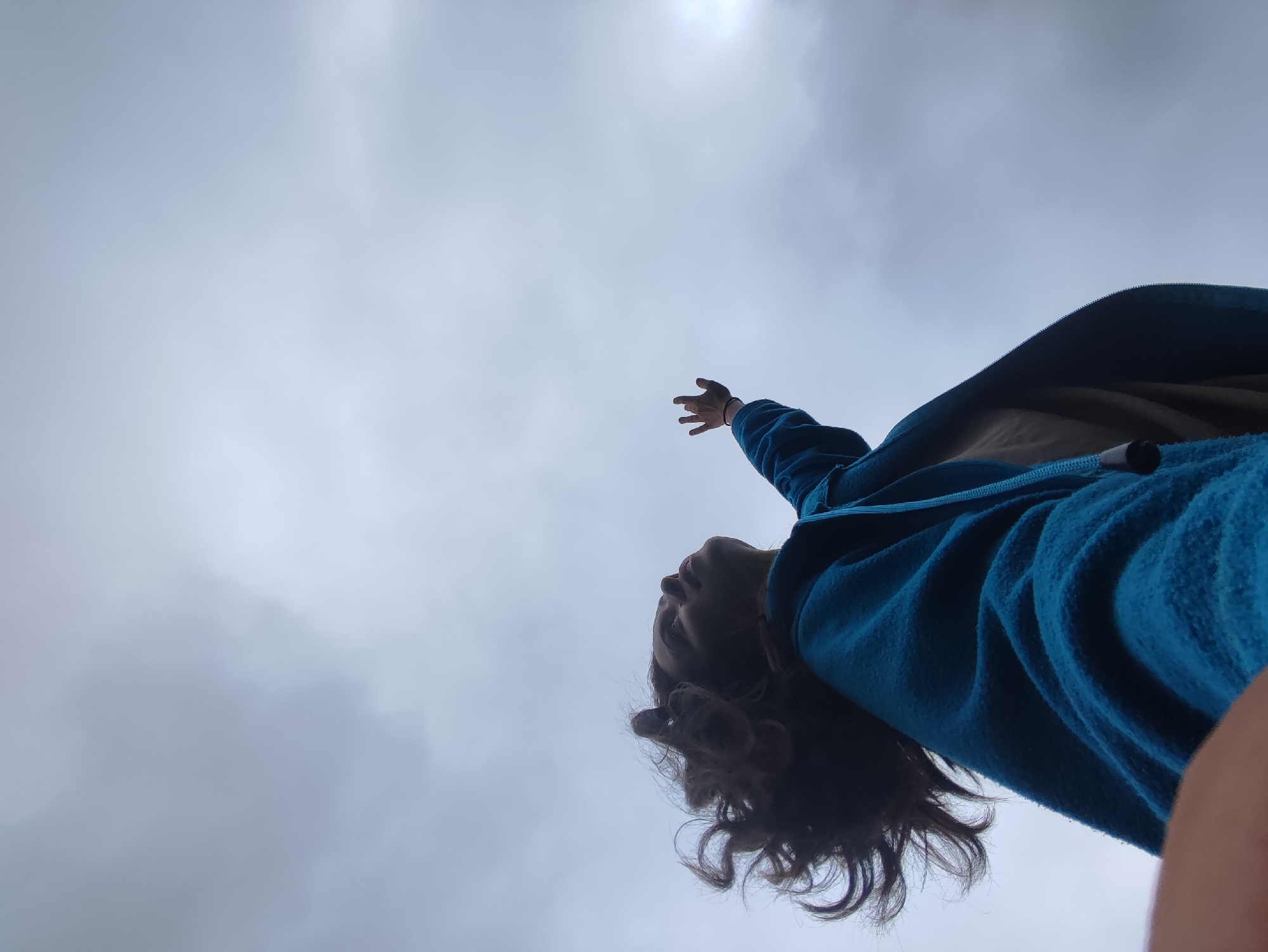 A low-angle, wide shot captures a person in a blue jacket reaching their left hand upwards into a cloudy sky. The person's head is tilted back, revealing their face and curly brown hair. The background is dominated by a gray, overcast sky with a hint of light in the upper right corner. The image appears to be taken outdoors, possibly in the city of Fokino, Russia, though this cannot be confirmed from the visual evidence alone.