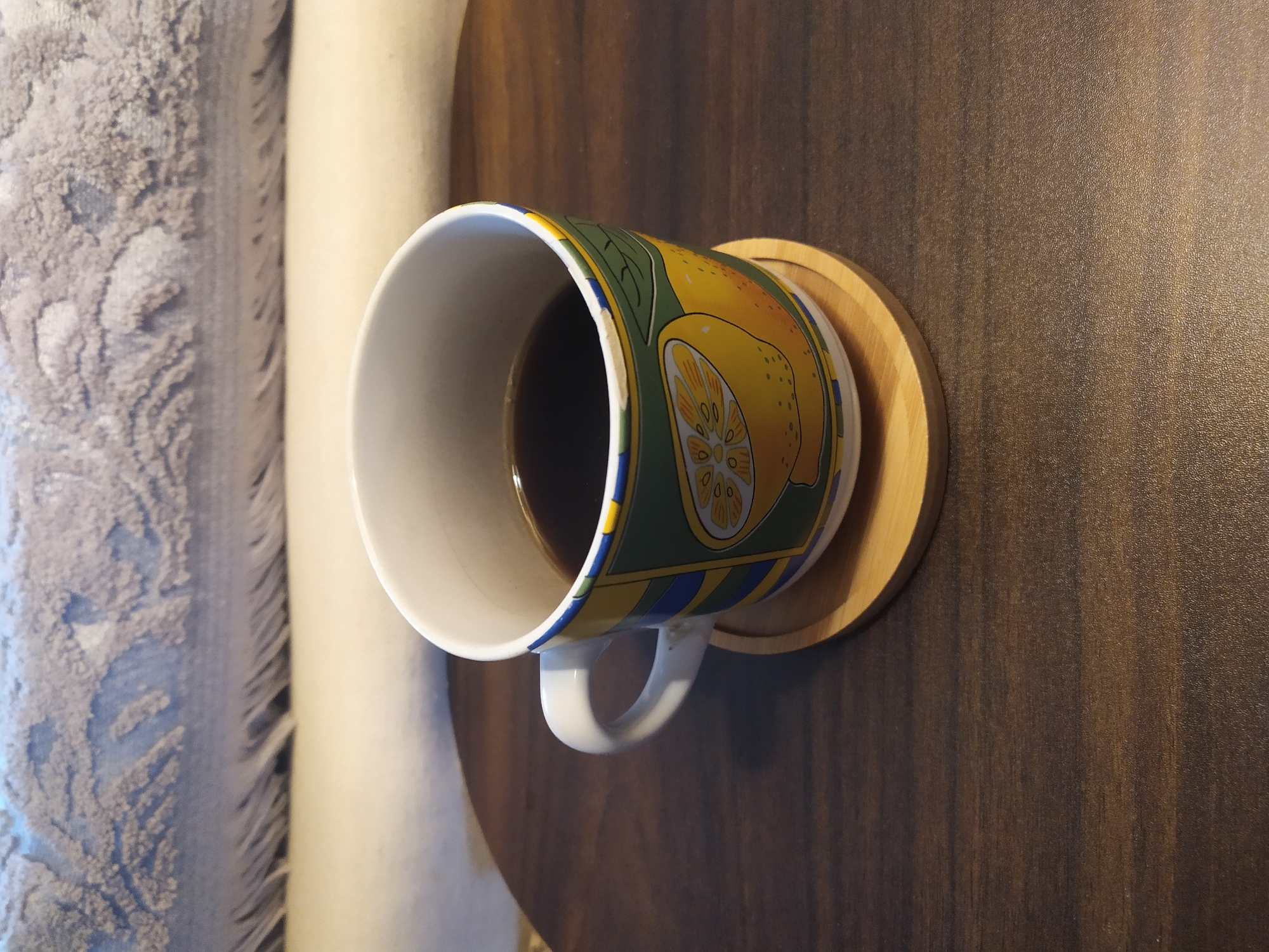 An overhead shot captures a colorful mug filled with dark coffee, resting on a round wooden coaster placed on a dark wooden table. The mug features a lively design of yellow lemons and green leaves. In the background, a plush, textured blanket with a fringed edge is draped horizontally, partially obscuring a light-colored surface beneath it. The lighting in the scene is soft and casts subtle shadows, creating a warm and inviting atmosphere.