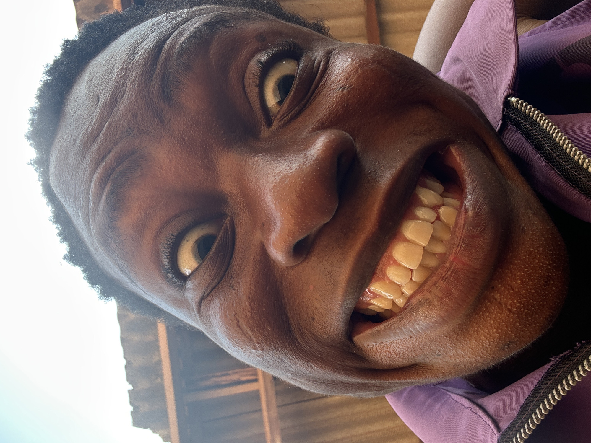 A close-up, low-angle, slightly rotated image shows a Black individual with dark skin, brown eyes, and short, curly hair. They are smiling broadly, displaying their teeth. The person is wearing a purple garment with a visible zipper on the right side of the image. The setting appears to be outdoors, with light from the sky visible in the top-left corner and the partial outline of what looks like a wooden structure in the background. Location is Unknown, Unknown.
