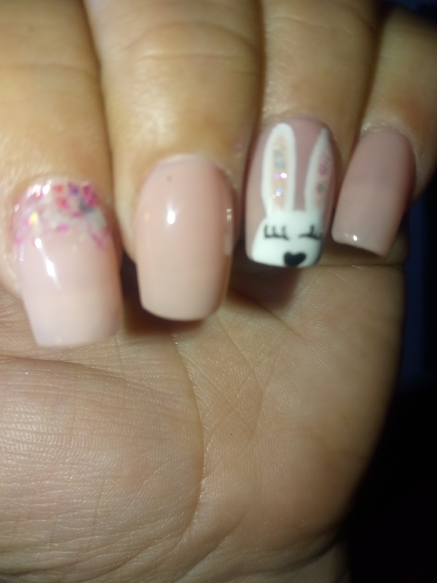 The image shows a close-up of a hand with decorated fingernails. The nails are painted in light pink or nude shades. One nail has a bunny face design with white ears, colorful glitter details, a black heart for a nose, and small lines for whiskers. Another nail features an arrangement of colorful glitter. The other nails are painted a solid color. The location and the city are unknown.