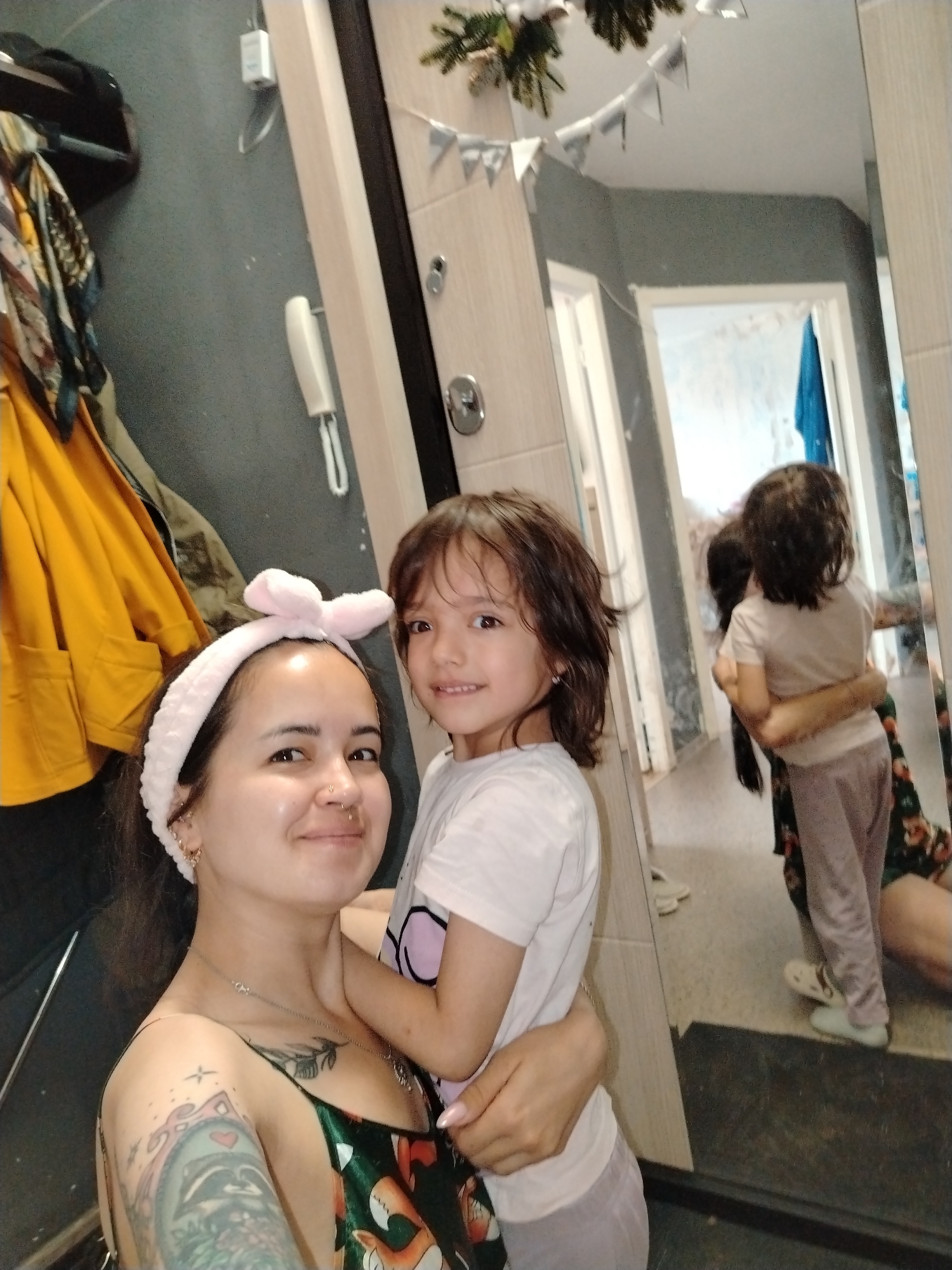 In the city of Noviy, Russia, a woman and a young child are reflected in a mirror, positioned in a home environment. The woman, wearing a pink headband with a bow, is in the foreground, holding the child. She has dark hair and is smiling, with visible tattoos on her arm and chest. The child, also with dark hair, is facing the camera, smiling and being held by the woman. They are both dressed casually.
The background, as seen in the mirror, reveals a hallway with a door and a tiled wall. Coats are hanging on a rack to the left of the woman, and a telephone receiver is mounted on the wall. Decorative elements, such as a string of triangular flags and greenery, are visible near the top of the mirror, suggesting a festive or decorative touch. The overall scene appears to be a candid moment captured within a domestic setting.