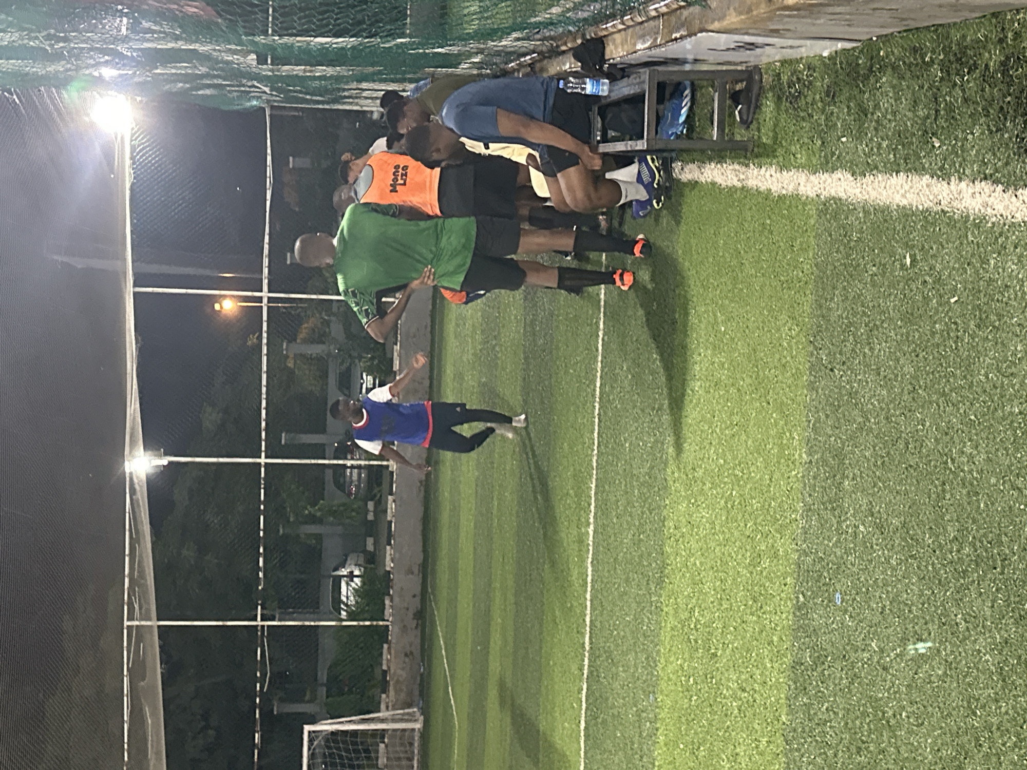 The image depicts an outdoor artificial turf soccer field at night, illuminated by artificial lighting. Several individuals are present on and around the field. In the foreground, a person wearing a blue and red bib over a white shirt and black shorts is walking on the field. Further back, a group of individuals are gathered, some standing and some sitting on a metal bench. One person in the group is wearing an orange bib with "Mona Liza" printed on it. Another person in a dark blue t-shirt and black shorts is seated on the bench. The background shows a fence, trees, and parked vehicles, suggesting an urban or semi-urban environment. The overall impression is of a casual soccer game or practice session. The provided context indicates the location is in Abuja, Nigeria.