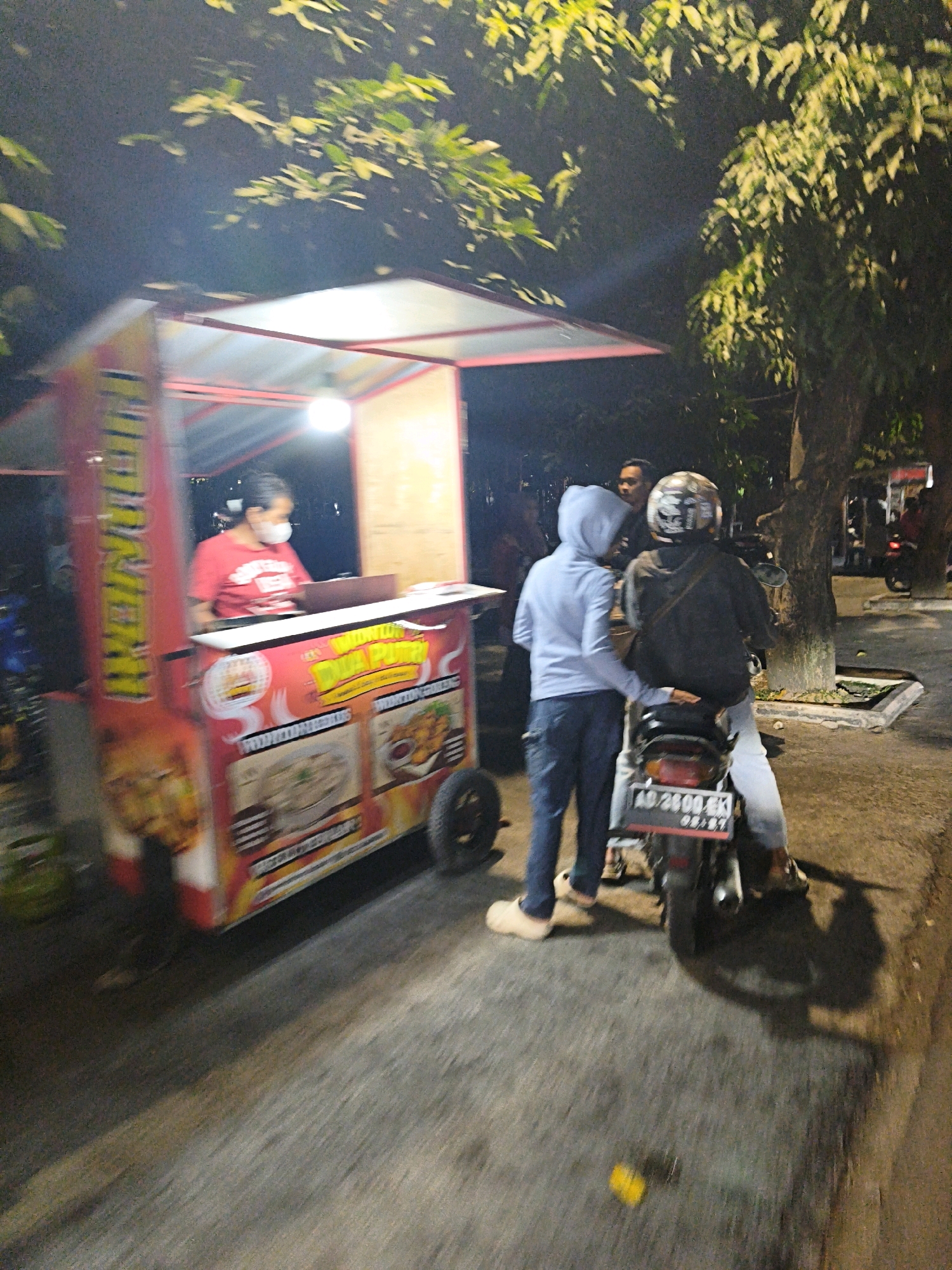 The image depicts a nighttime scene on a street in Surakarta, Indonesia, near a food cart. A person wearing a red shirt and a white face mask is behind the counter of the food cart, which is illuminated by a bright light. The cart is decorated with colorful images of food. In front of the cart, two individuals are visible. One, wearing a light blue hoodie and jeans, is standing next to a motorcycle. The other, wearing a dark jacket and a helmet, is seated on the motorcycle, which has a license plate that reads "AD 2800 EK". The background features trees and the dark night sky. The street appears to be asphalt. The overall lighting suggests it is nighttime, with artificial lights from the food cart and possibly streetlights illuminating the area.