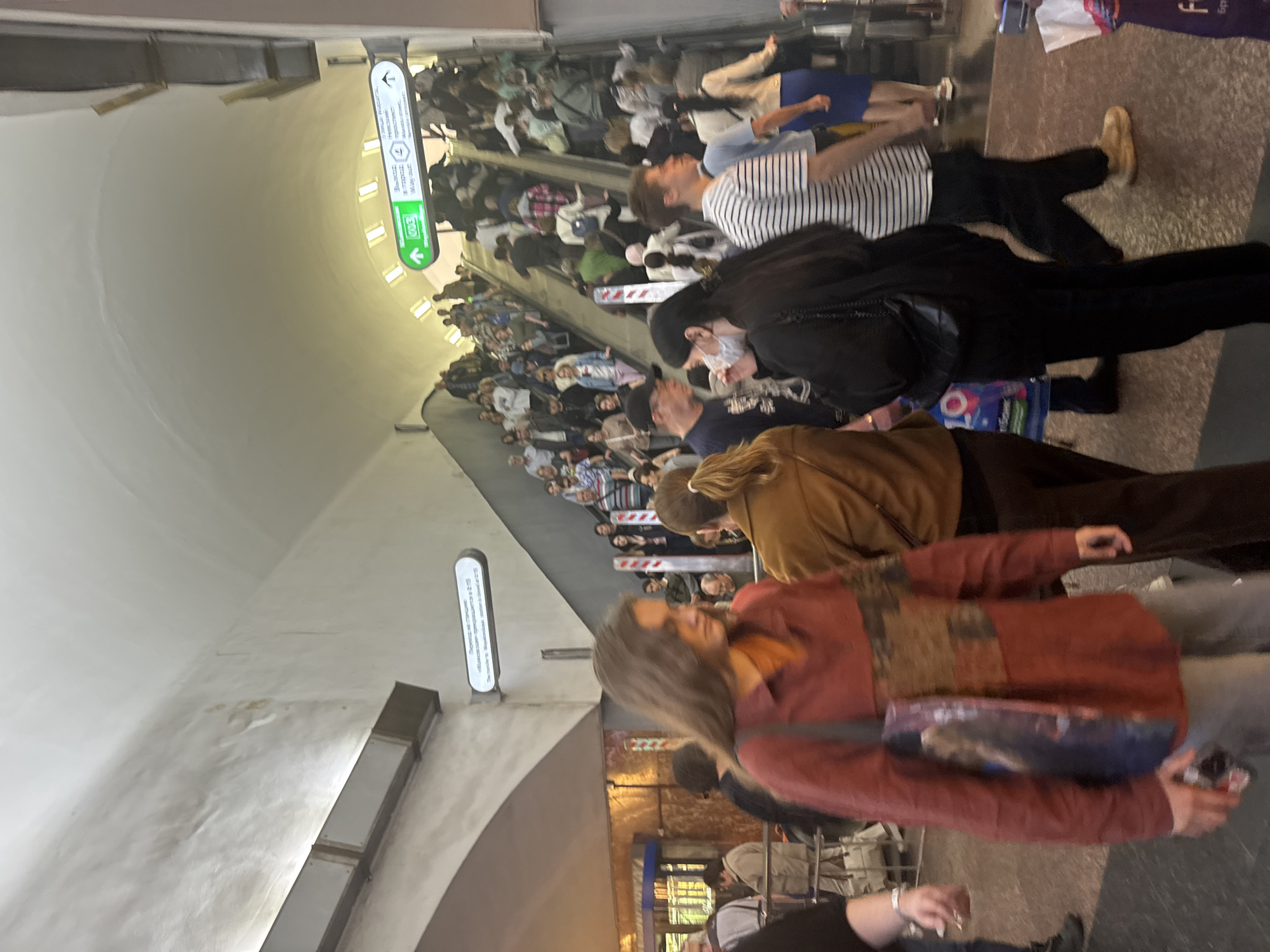 The image depicts an interior scene, likely a subway station in Saint Petersburg, Russia, characterized by a large, arched ceiling and escalators filled with people. The environment appears to be an underground passageway.
Several individuals are visible, predominantly moving on escalators or standing in the vicinity. One person in the foreground, on the left, is wearing a reddish-brown long-sleeved shirt and carrying a tote bag. To their right, a woman with long brown hair is looking down. Further to the right, a woman in a brown jacket is visible.
Above the escalators, there are directional signs. One sign, illuminated in green, points upwards and to the right, with Cyrillic text and numbers indicating "4" and "2:15". Another sign to its left also has Cyrillic text. The escalators themselves are populated with a diverse group of people, some ascending and others descending, engaged in their commute.
The lighting in the scene is a mix of artificial light from the ceiling fixtures and the illuminated signs. The overall impression is one of a busy public transportation hub.