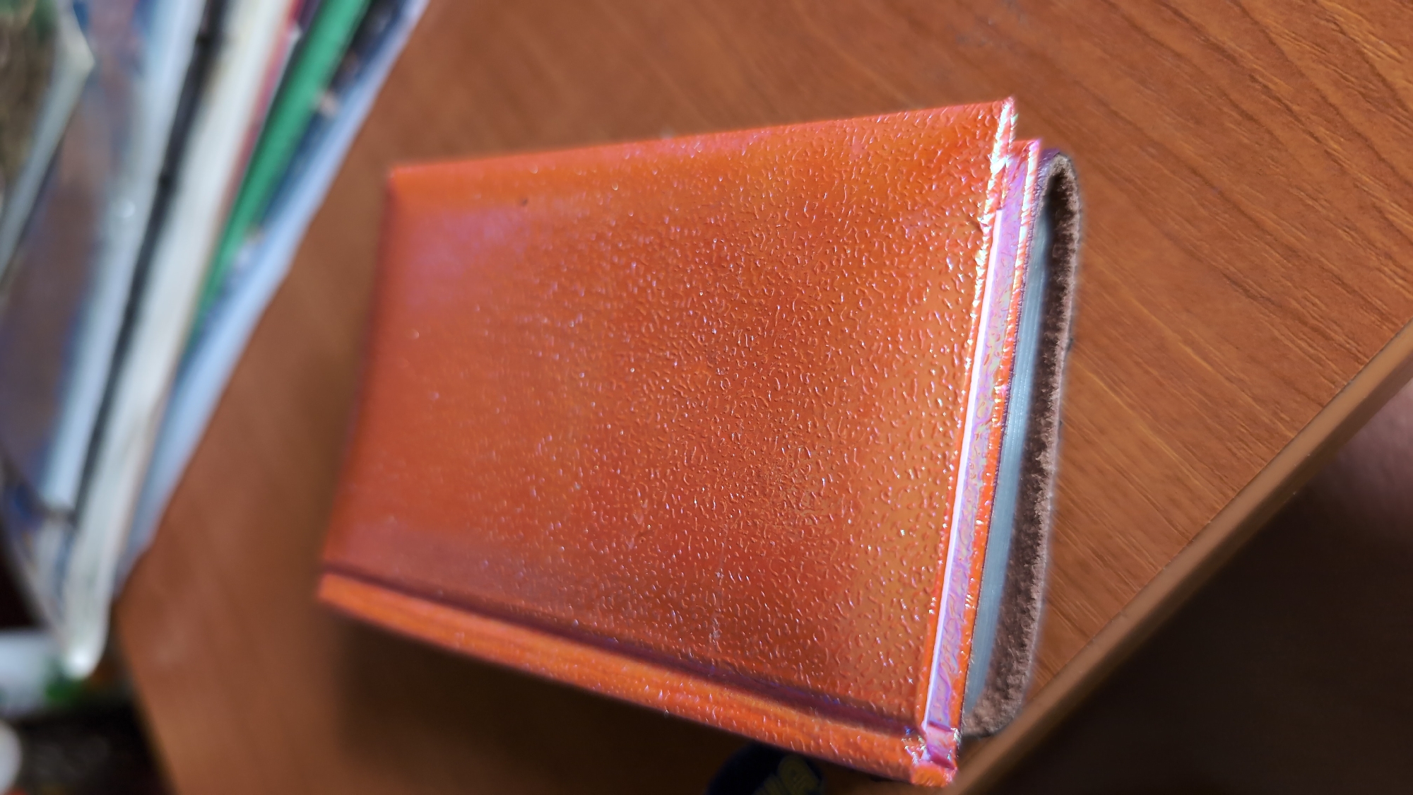 The image shows a close-up, angled view of an orange, textured object lying on a wooden surface. The object appears to be a small book or notebook, with its spine visible on the right side. The spine is covered in a brown, fuzzy material, possibly leather or suede, and there are visible pages, which are light blue. The orange cover has a subtle, iridescent sheen, reflecting light in shades of pink and purple. The texture of the cover is small and repetitive, like tiny bumps or dimples.
To the left of the orange object, a stack of other items is visible, out of focus. These items appear to be books or binders with spines of various colors, including white, blue, and green. The overall setting suggests an indoor environment, likely a desk or table, given the wooden surface and the presence of books. There is no indication of people, a specific time of day, or weather conditions. The scene is static, with no activity taking place. No visible text is present on the orange object or the surrounding items. The location context is Mykolaiv, Ukraine.