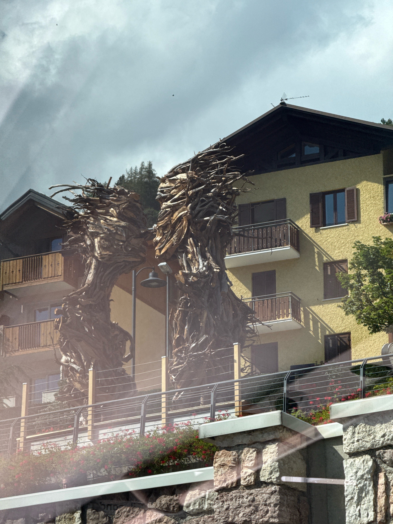 The image depicts a large, abstract sculpture made of wood, resembling intertwined branches or roots, situated in front of a building in Molveno, Italy. The sculpture is positioned on a tiered terrace with metal railings and blooming red flowers. The building behind it is yellow with dark brown shutters and balconies. In the background, a forest of coniferous trees is visible under a cloudy sky. The sculpture appears to be the main subject, with the building and natural surroundings serving as its context.