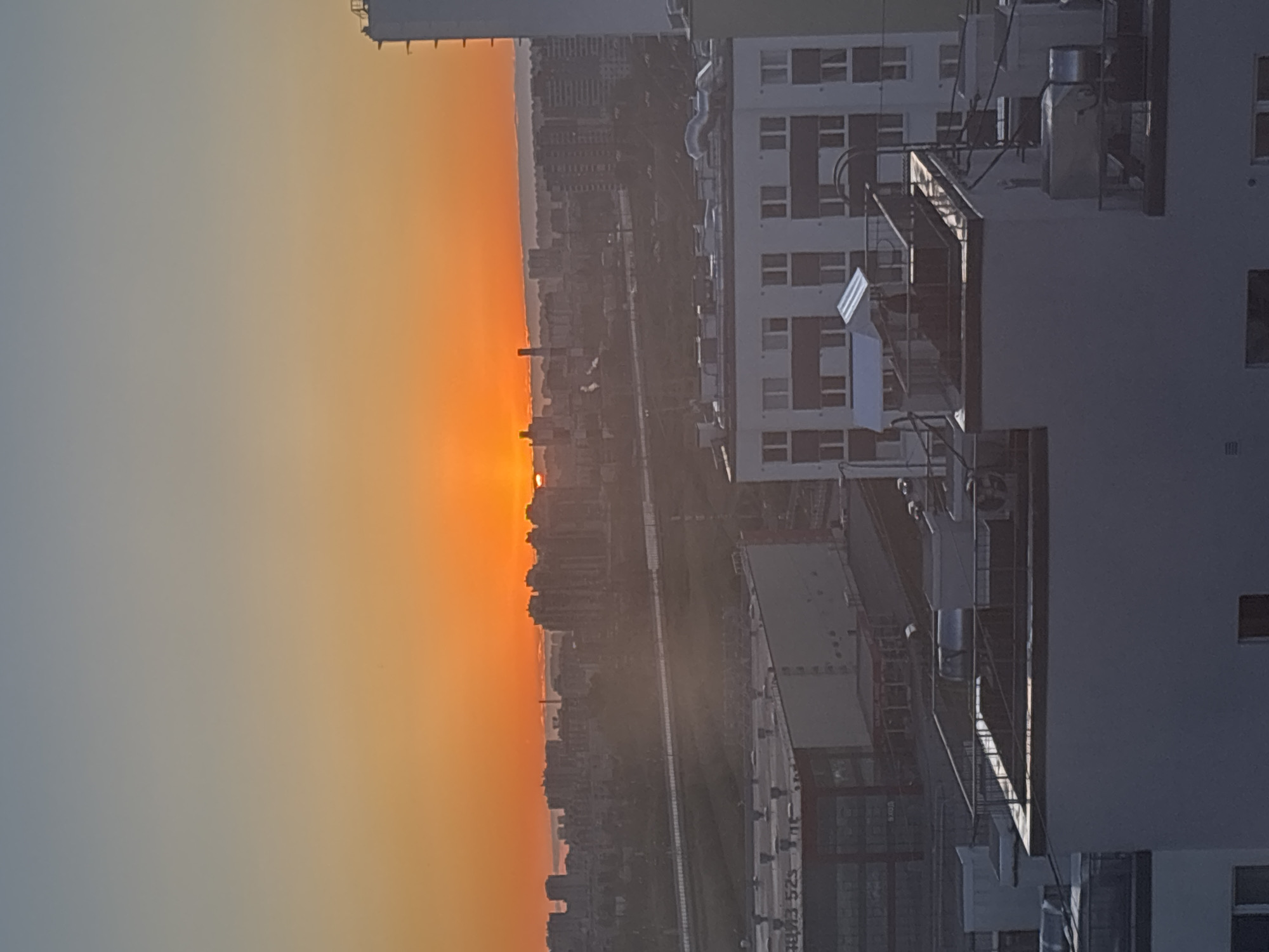 The image shows a sunset over Vsevolozhsk, Russia. The sky is orange and yellow where the sun is setting over the urban area. Tall buildings and cityscape structures are visible silhouetted against the bright sky. A building with multiple windows and balconies is in the foreground. Some equipment is mounted on the building. A train track is also visible in the middle ground.